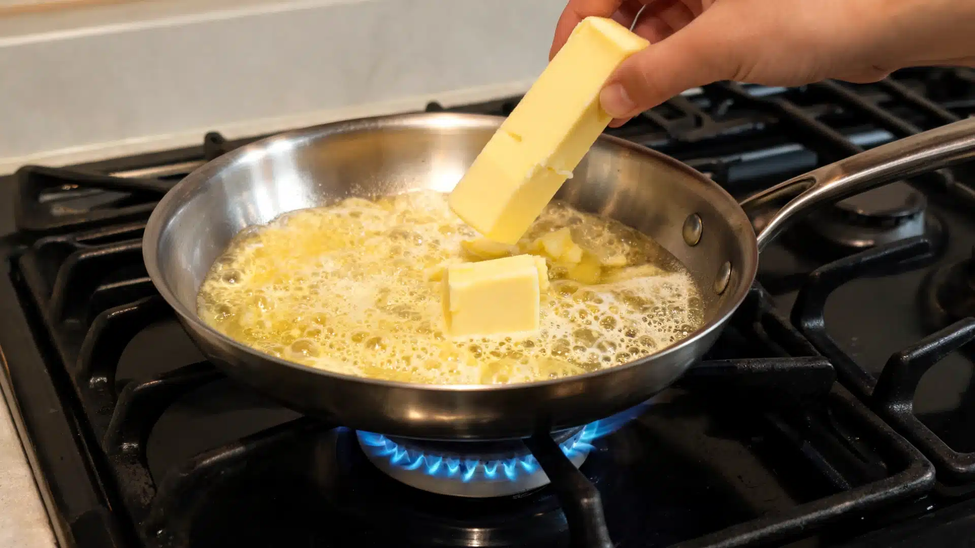 hand adding butter to a pan of melting butter on a gas stove while it bubbles over low heat