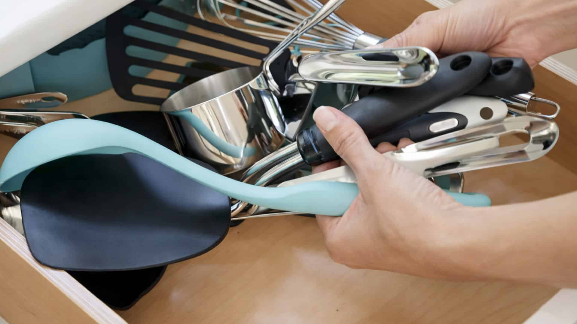 hands removing mixed kitchen utensils in an open drawer, including spatula, whisk, measuring cup, and tongs.