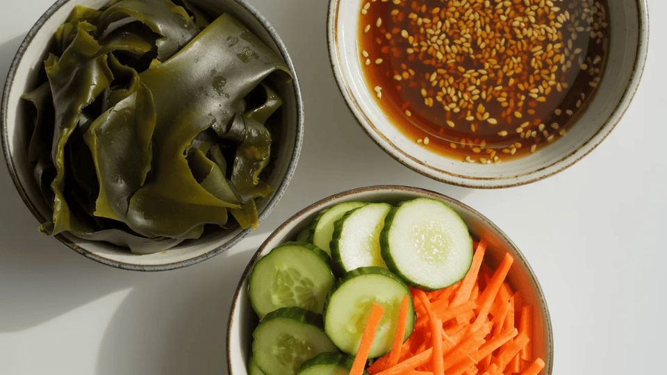 overhead view of three bowls with rehydrated wakame seaweed, sliced cucumber with shredded carrots, and sesame soy dressing