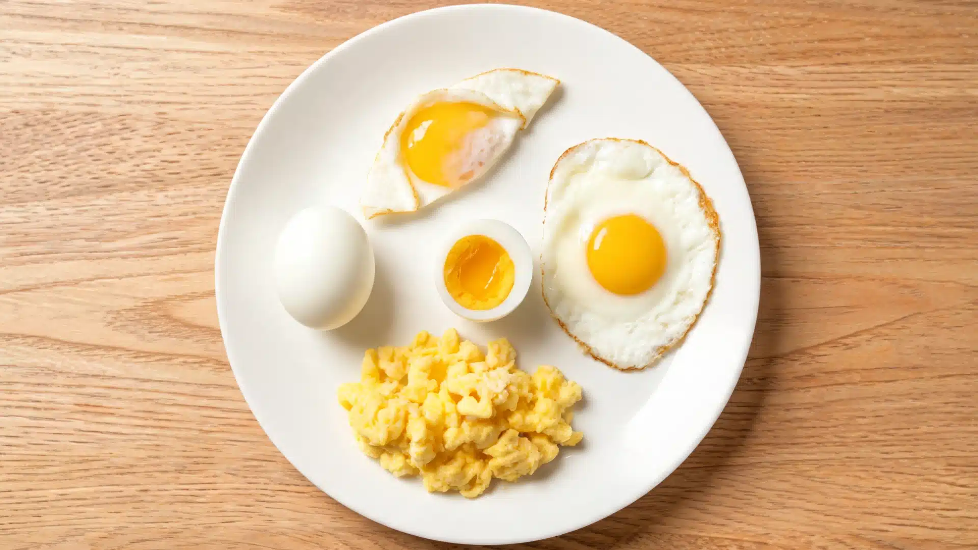 plate of assorted cooked eggs including fried egg, soft boiled egg, hard boiled egg, and scrambled eggs served on a wooden table in natural light