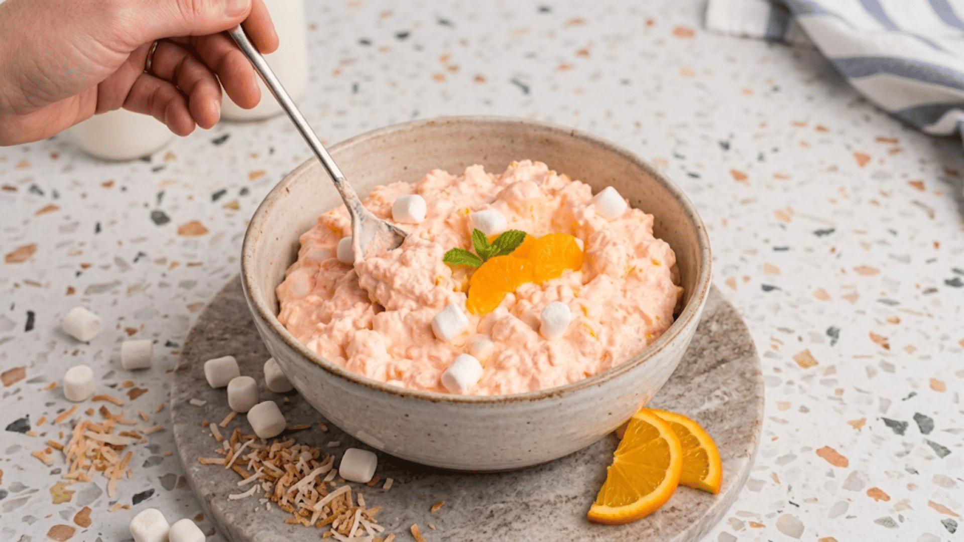 a bowl of creamy orange gelatin fluff topped with mandarin slices marshmallows and mint with a spoon scooping a serving