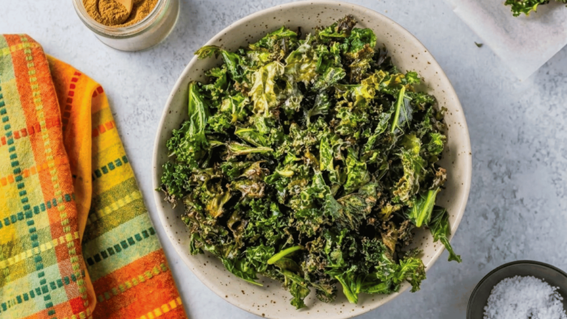 a bowl of crispy kale chips seasoned with spices served on a table beside sea salt and a colorful kitchen towel for snacking