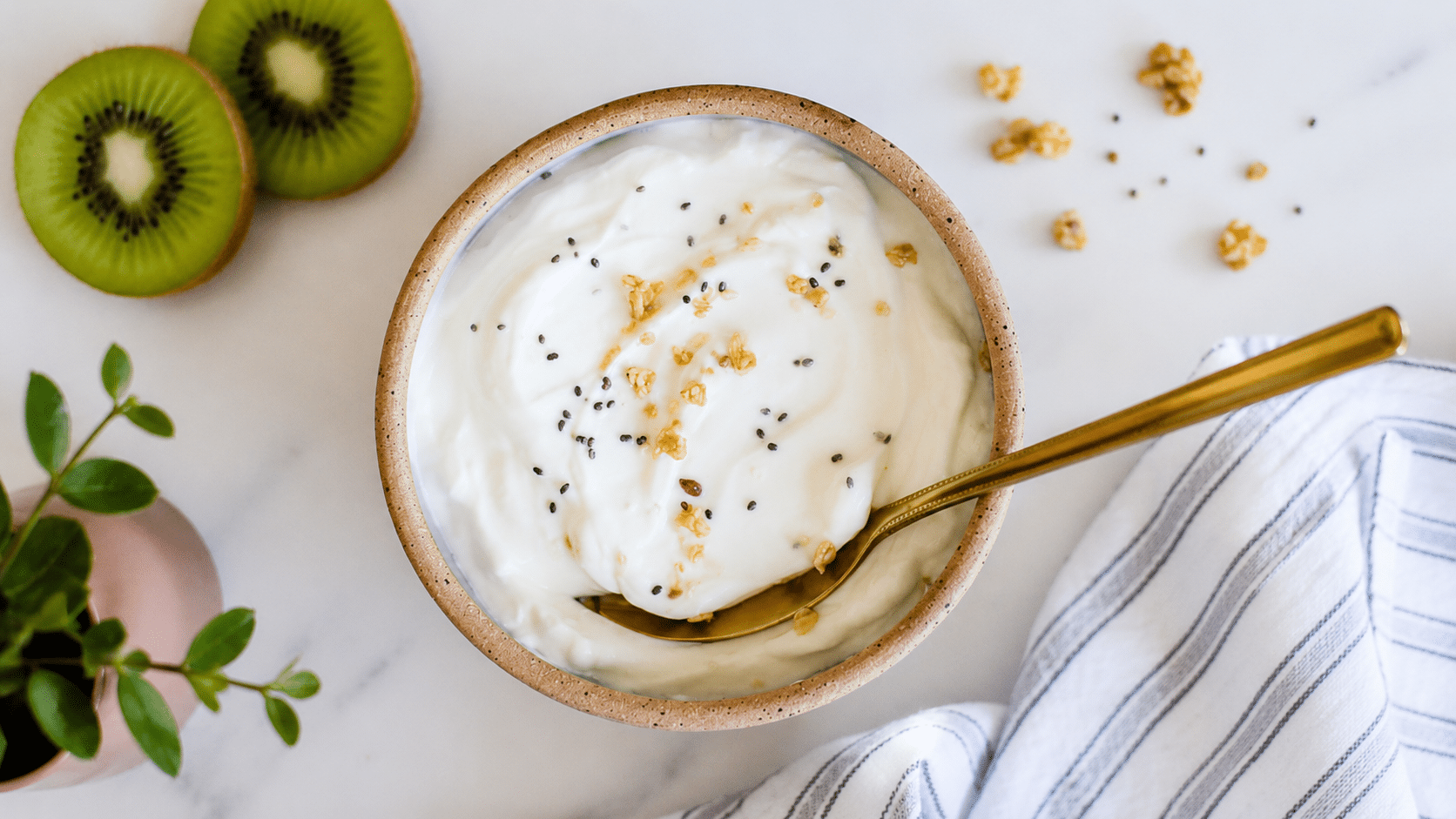 a bowl of greek yogurt topped with chia seeds and granola beside sliced kiwi and a gold spoon on a white marble surface