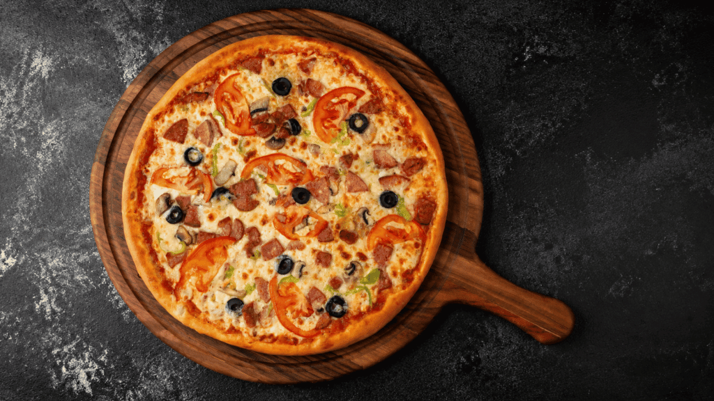 a freshly baked pizza topped with tomatoes olives mushrooms ham and melted cheese on a wooden board over a dark surface