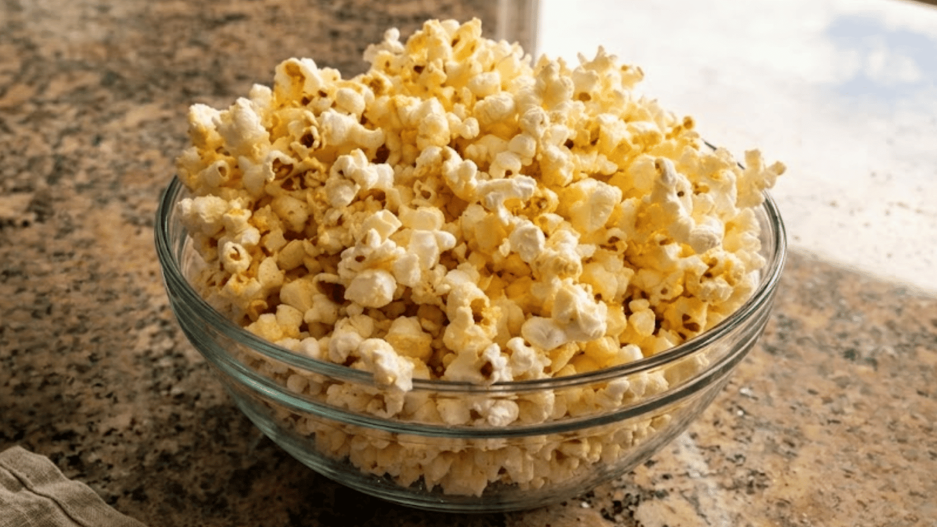 a glass bowl filled with freshly popped popcorn sitting on a granite countertop beside a bright window ready to enjoy