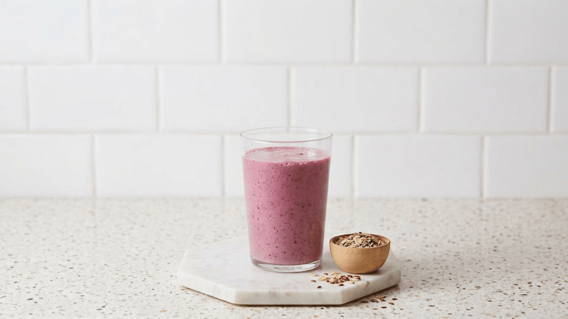 a pink berry gelatin smoothie in a clear glass beside a small bowl of oats on a marble board against a white tiled background