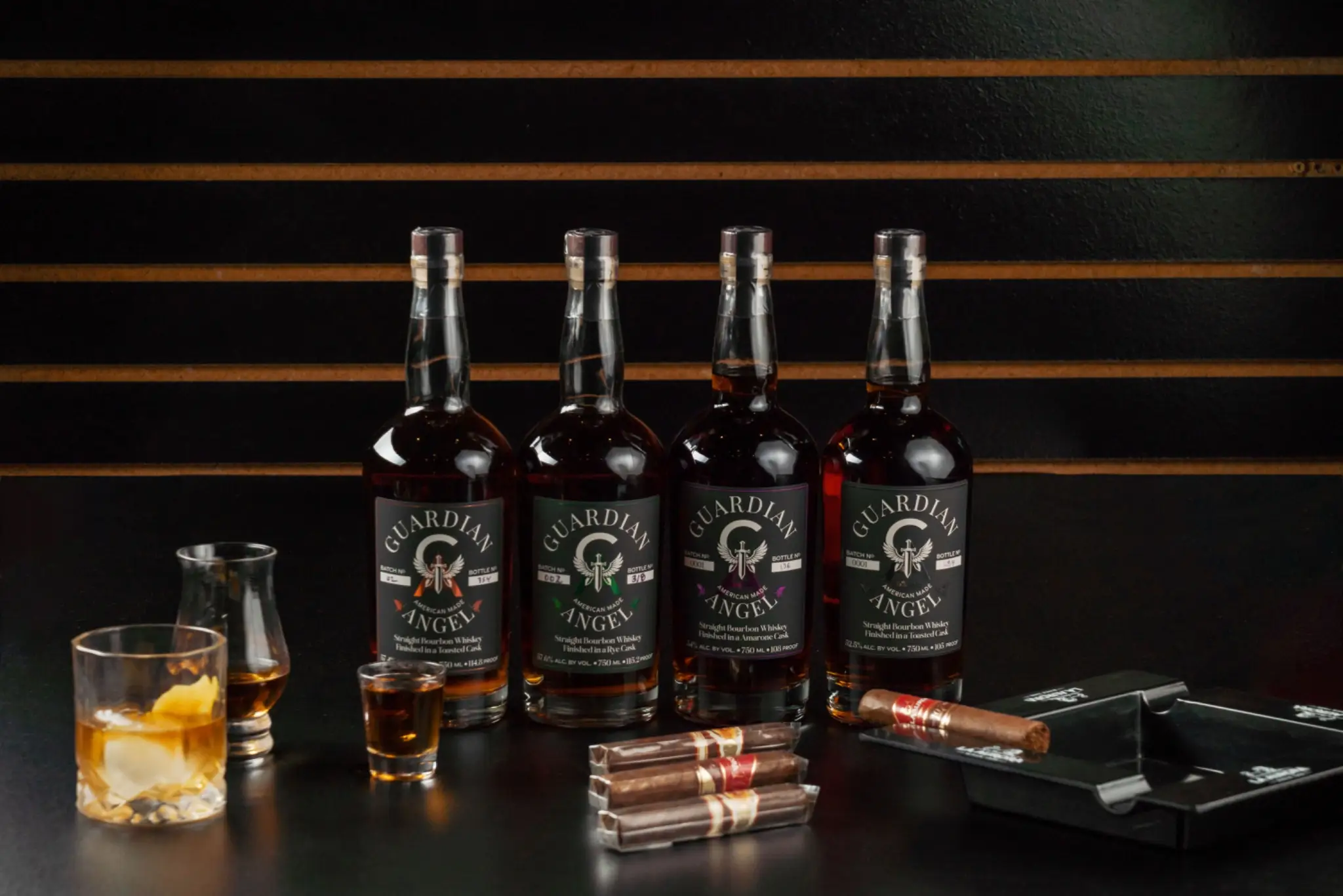 Four bottles of bourbon with cigars and whiskey glasses on dark tabletop