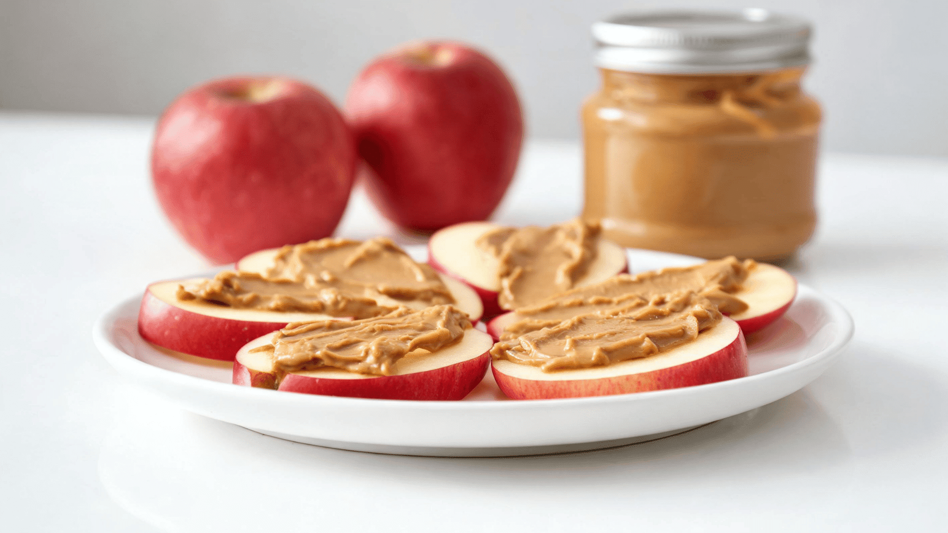 apple slices topped with peanut butter on a white plate beside whole apples and a jar of peanut butter