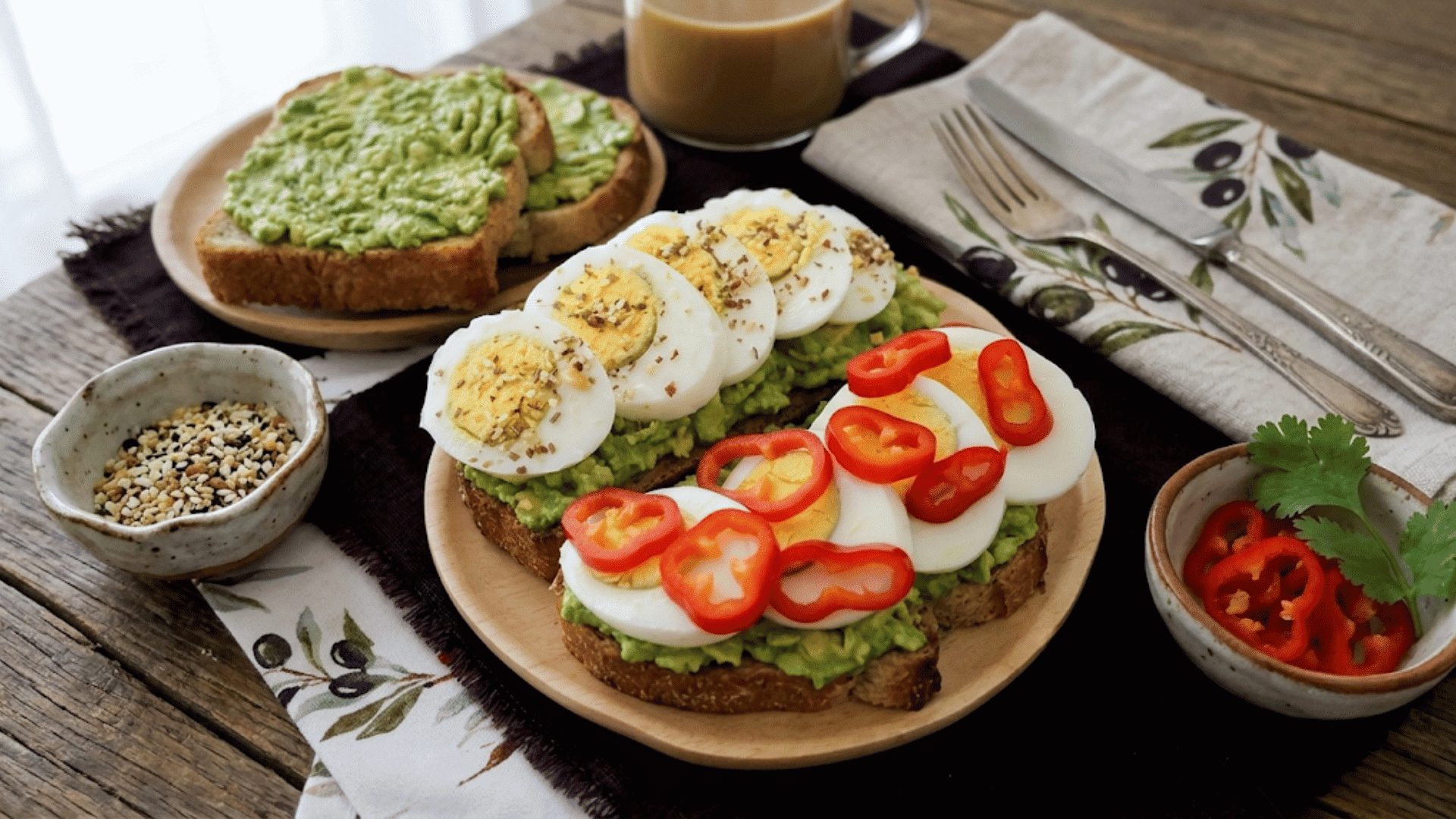 avocado toast topped with sliced hard boiled eggs red pepper rings and seasoning served on a wooden plate with toast in background (2)