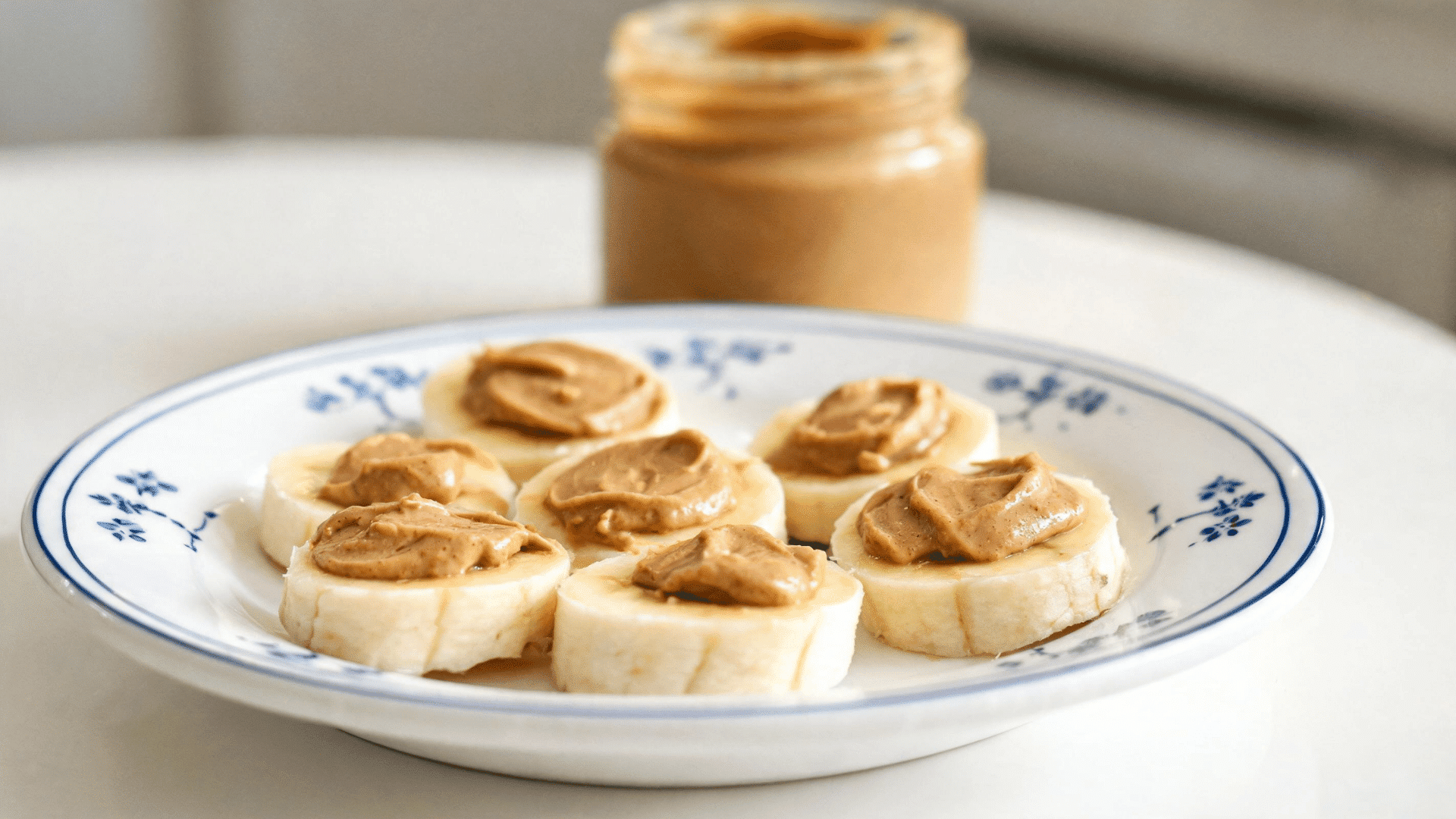 banana slices topped with almond butter on a patterned plate beside an open jar of peanut butter