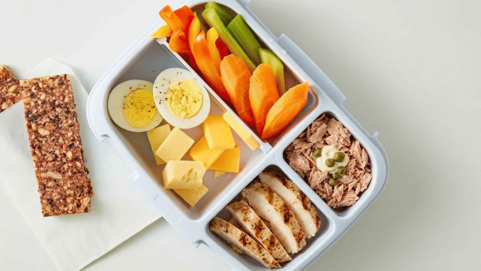 bento lunch box with boiled eggs cheese grilled chicken tuna sliced vegetables and granola bar arranged in compartments