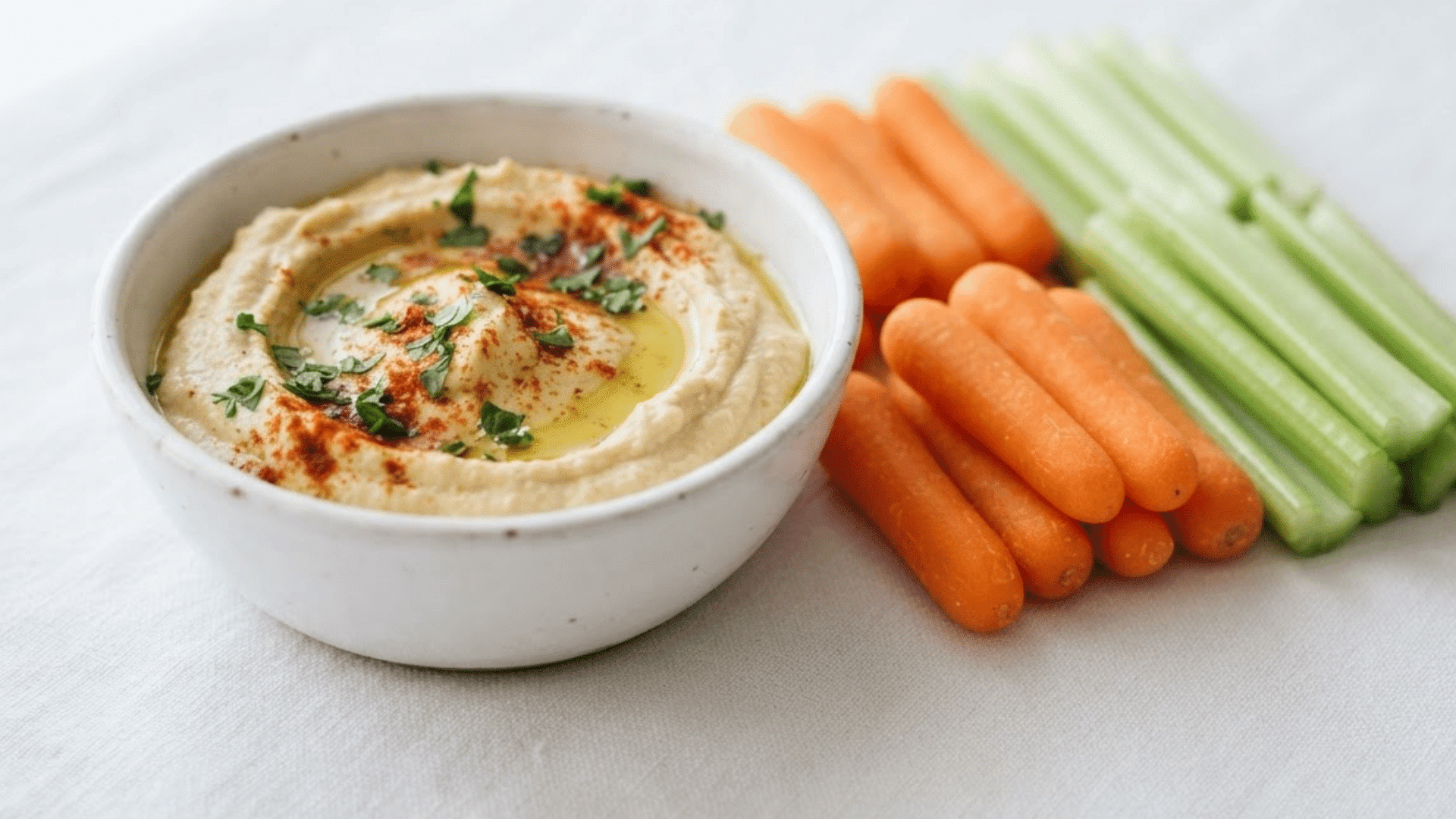 bowl of creamy hummus with baby carrots and celery sticks arranged on a clean white surface in natural light