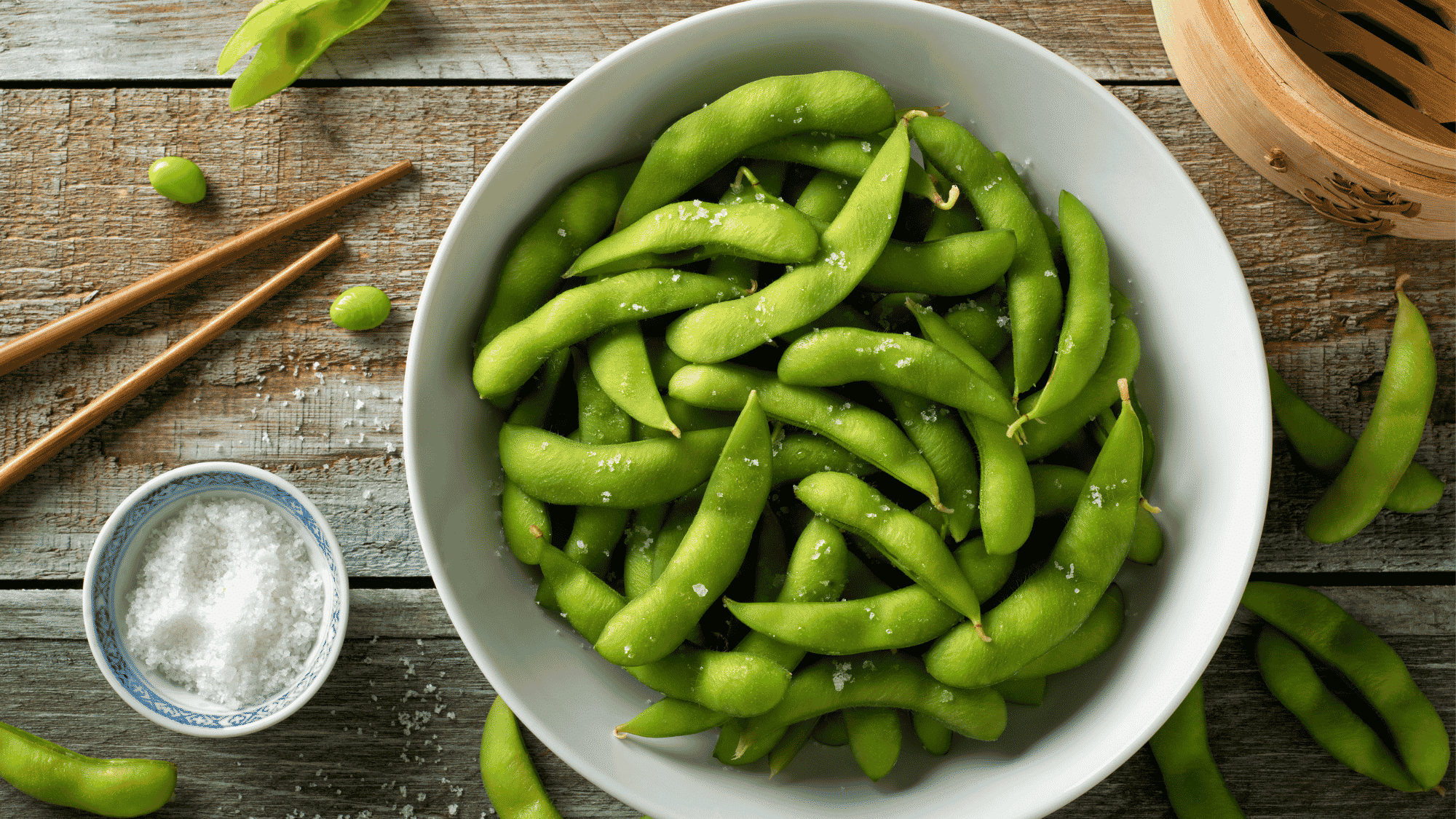 bowl of fresh edamame beans with sea salt on rustic wooden table with chopsticks and dipping bowl