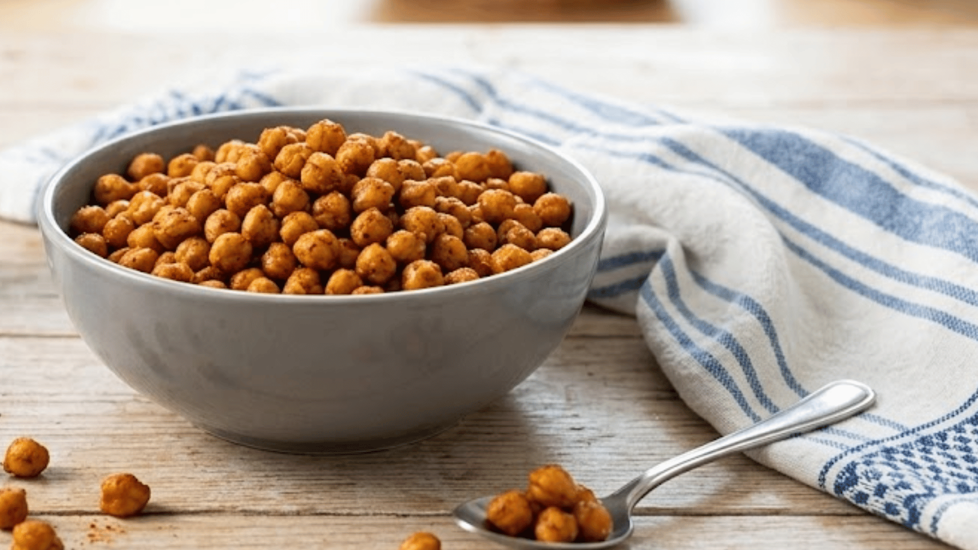 bowl of roasted chickpeas seasoned with paprika served on wooden table beside salt spices and spoon with kitchen background