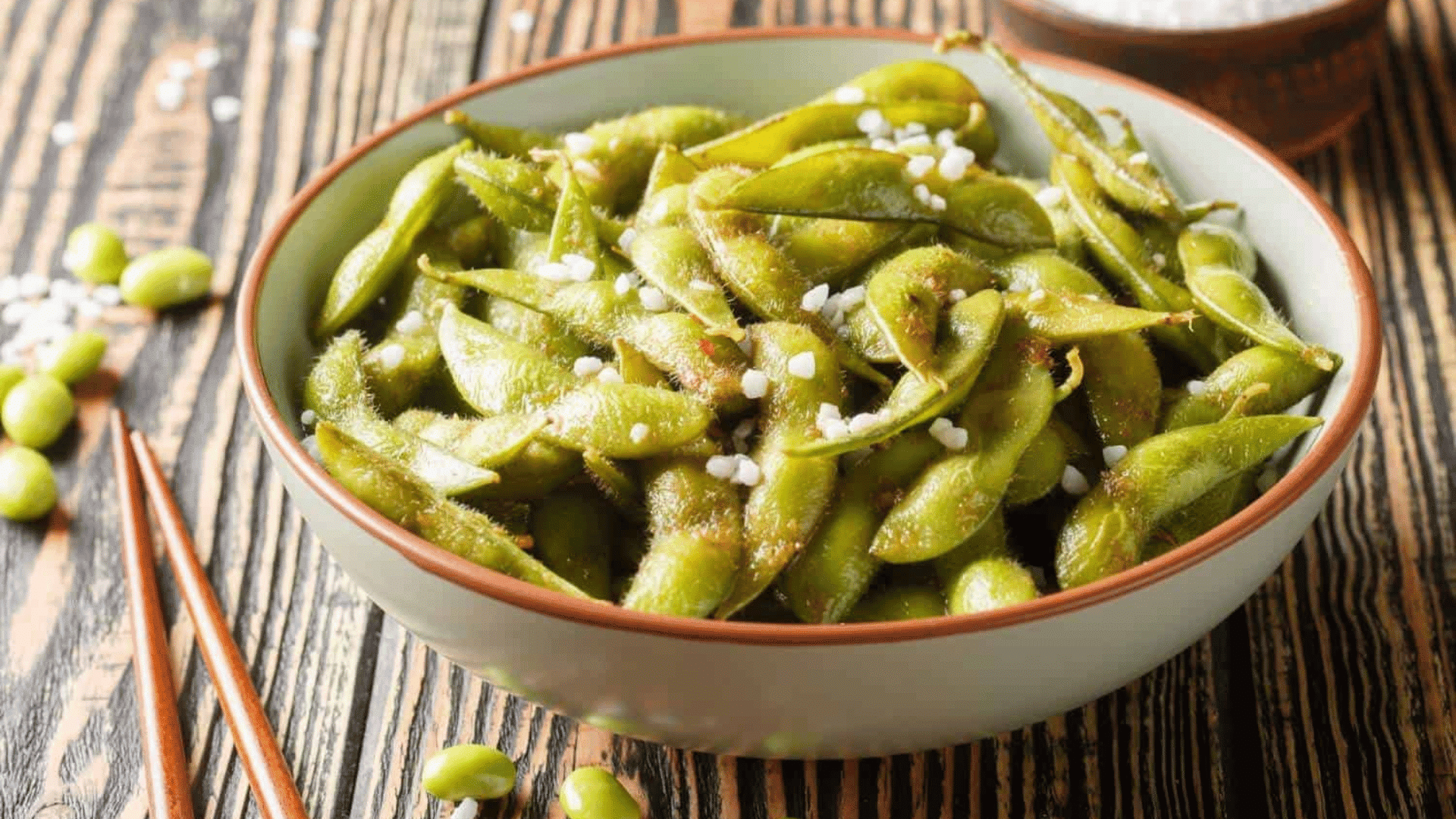 bowl of steamed edamame sprinkled with coarse salt served on wooden table beside chopsticks and loose soybeans