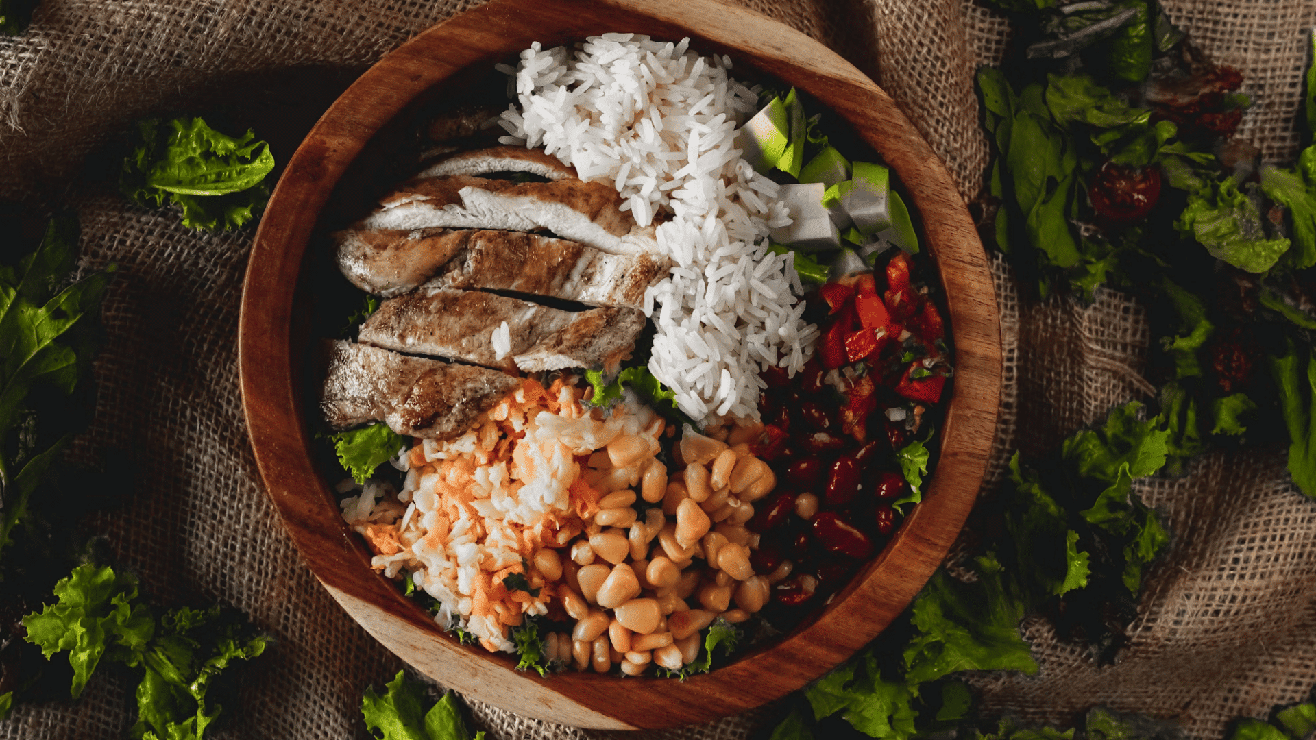 bowl with grilled chicken white rice vegetables and beans arranged on a rustic cloth surface