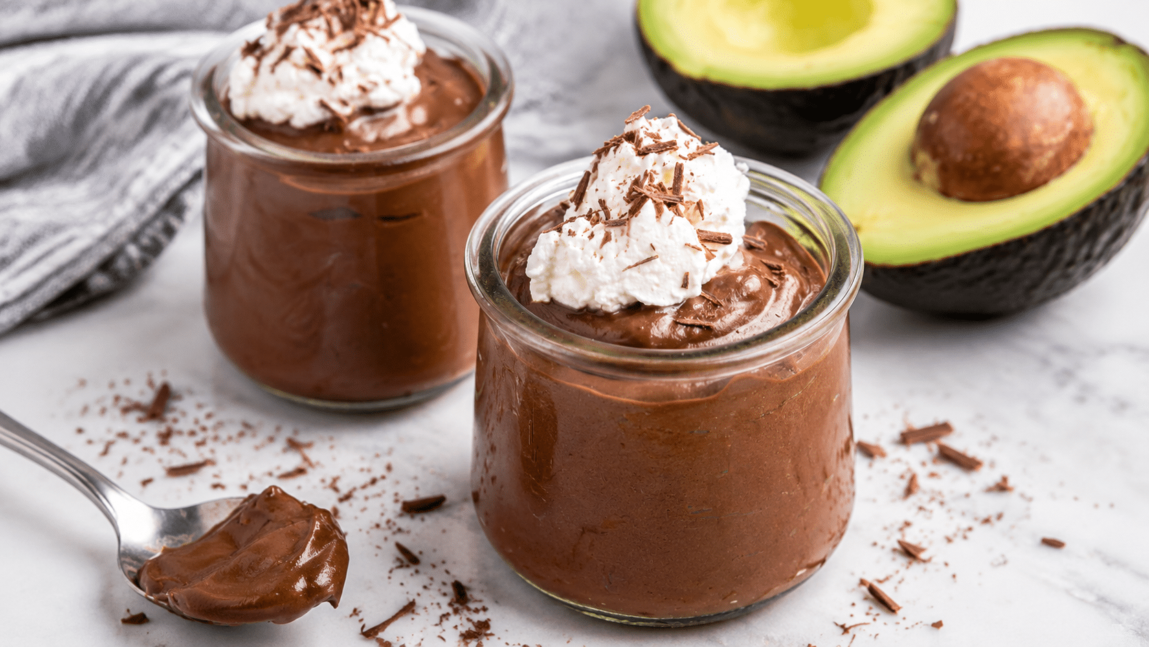 creamy avocado chocolate mousse topped with whipped cream and chocolate shavings served in glass jars with avocado halves