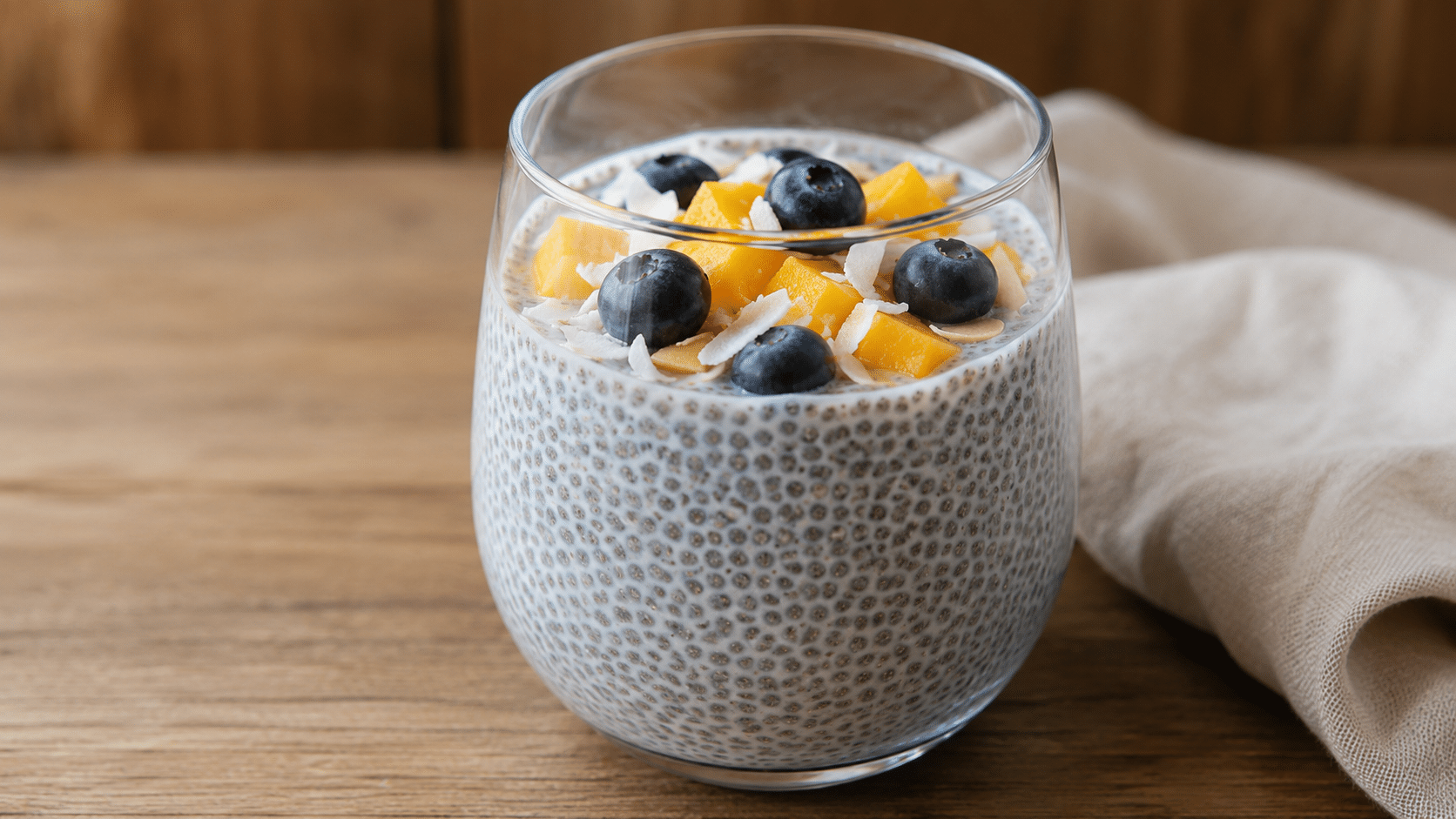 creamy chia seed pudding topped with fresh mango blueberries coconut flakes and sliced almonds in a glass on a rustic wooden table
