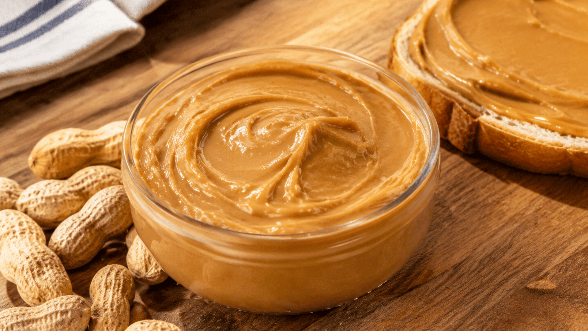creamy peanut butter in a glass bowl with whole peanuts and peanut butter spread on bread over a wooden kitchen surface.