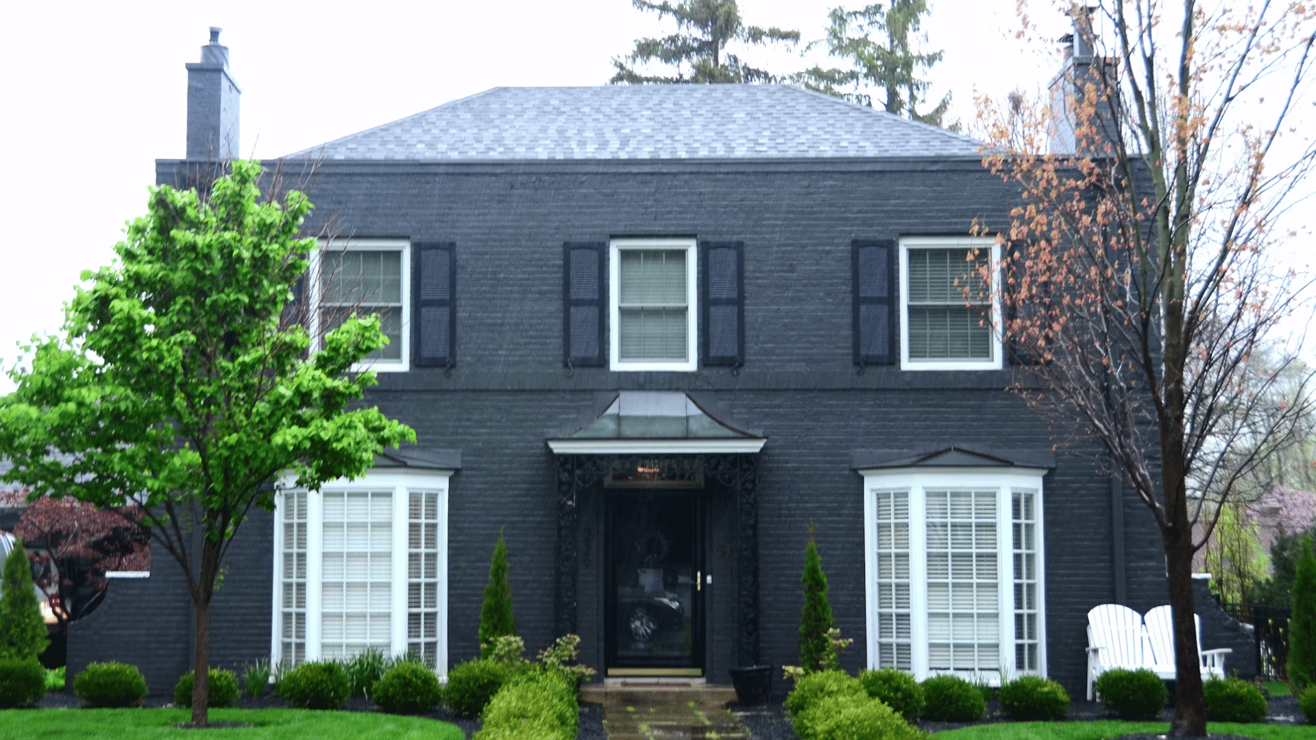 dark brick house with white trim windows green lawn and shrubs along a front pathway under cloudy sky