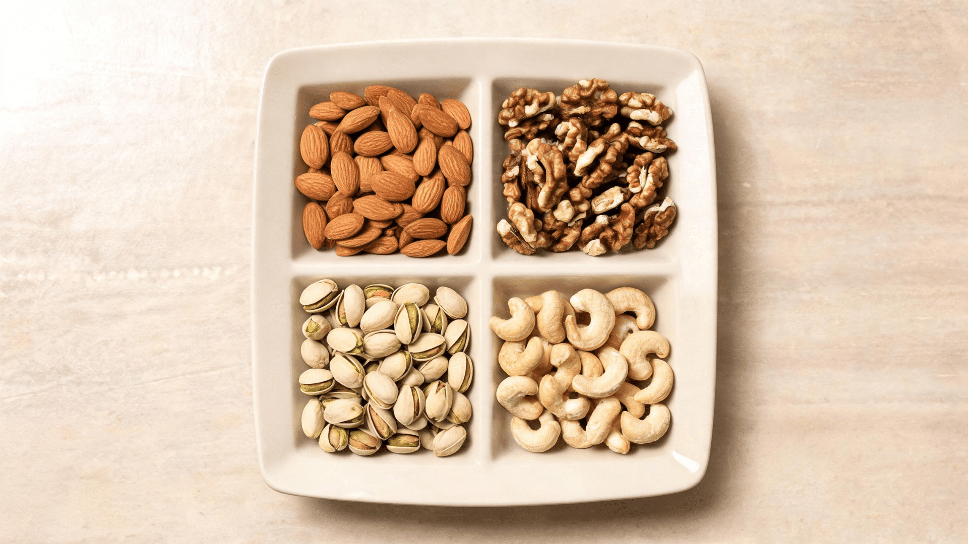 Four section plate filled with almonds walnuts pistachios and cashews on a light stone surface