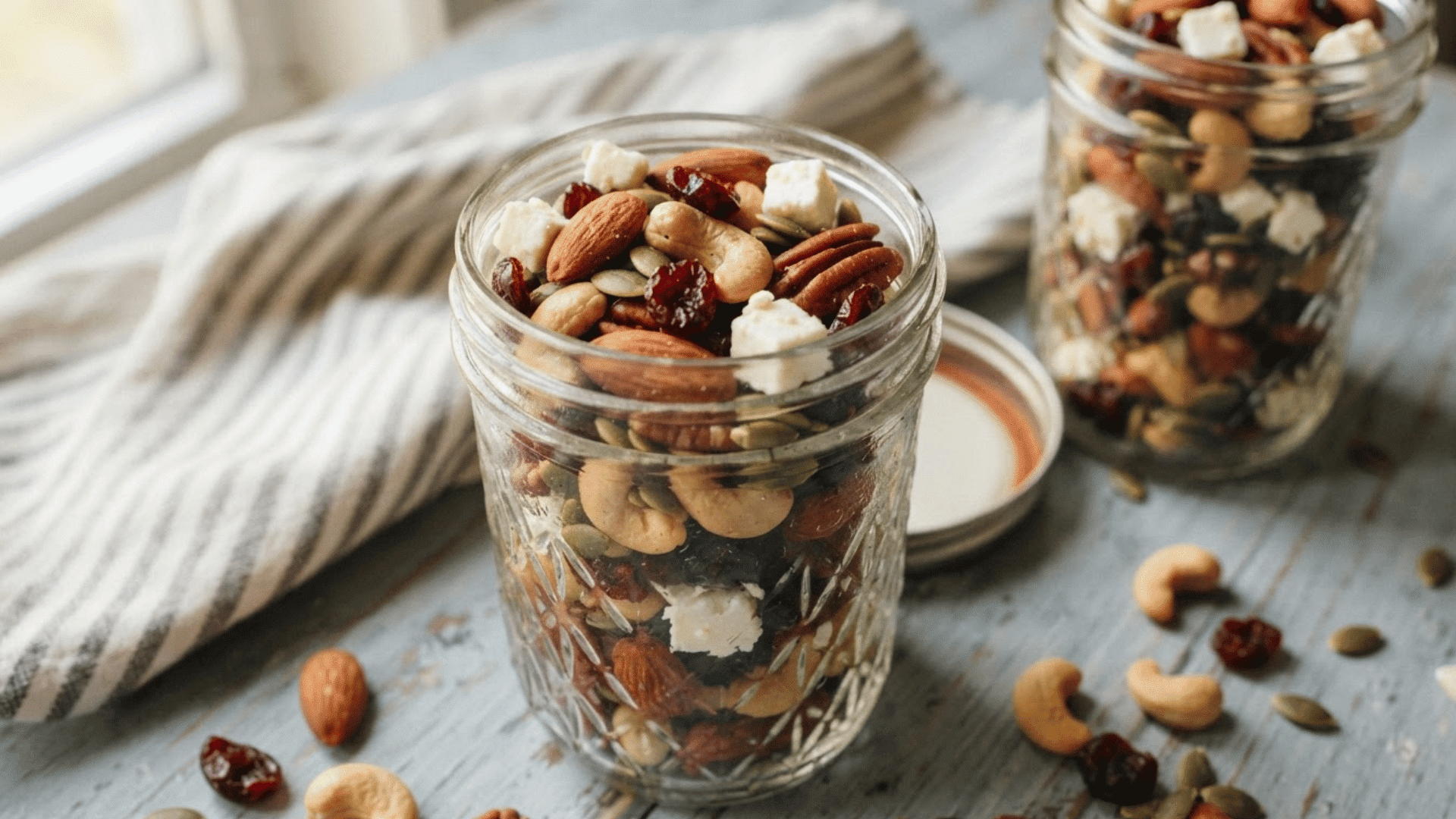glass jar filled with mixed nuts seeds dried fruits and cheese pieces on a light surface with a second jar behind