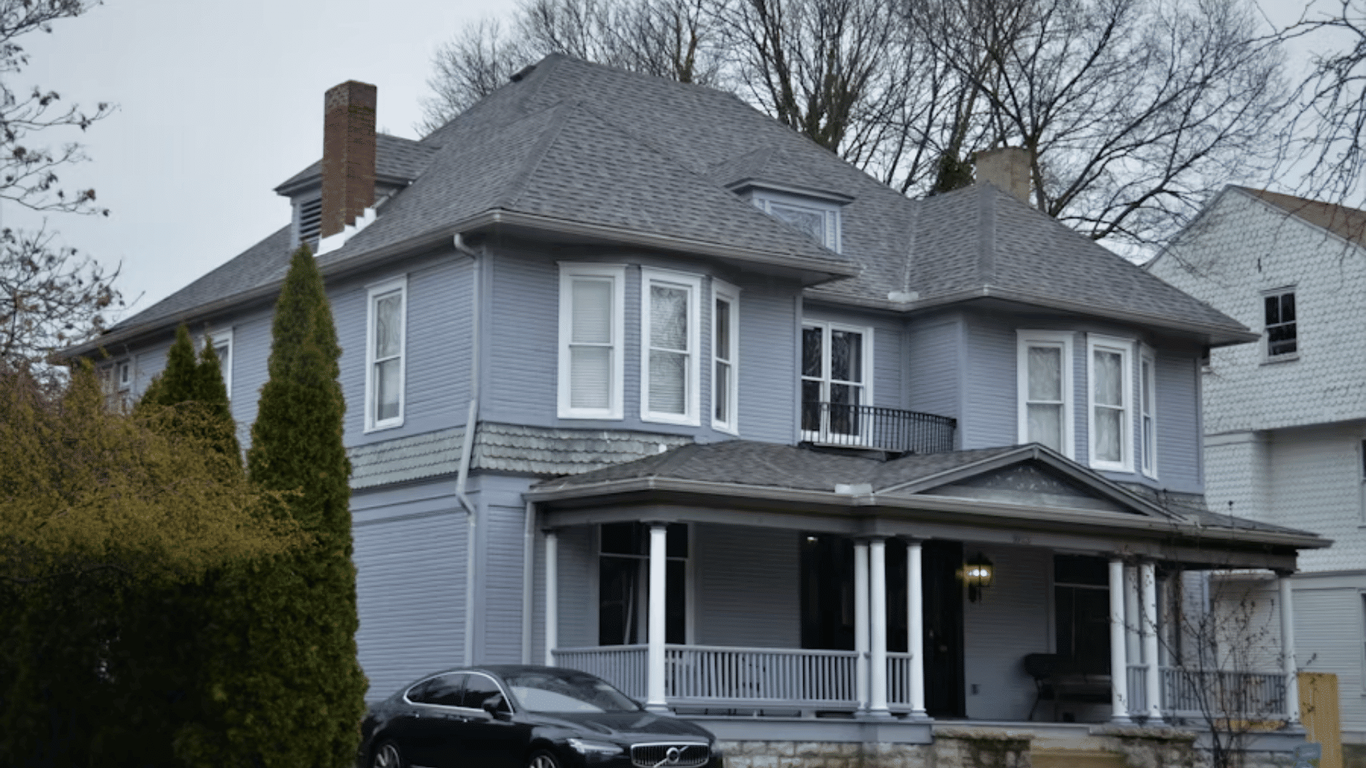 gray two story house with covered porch balcony and a black car parked beside the front lawn