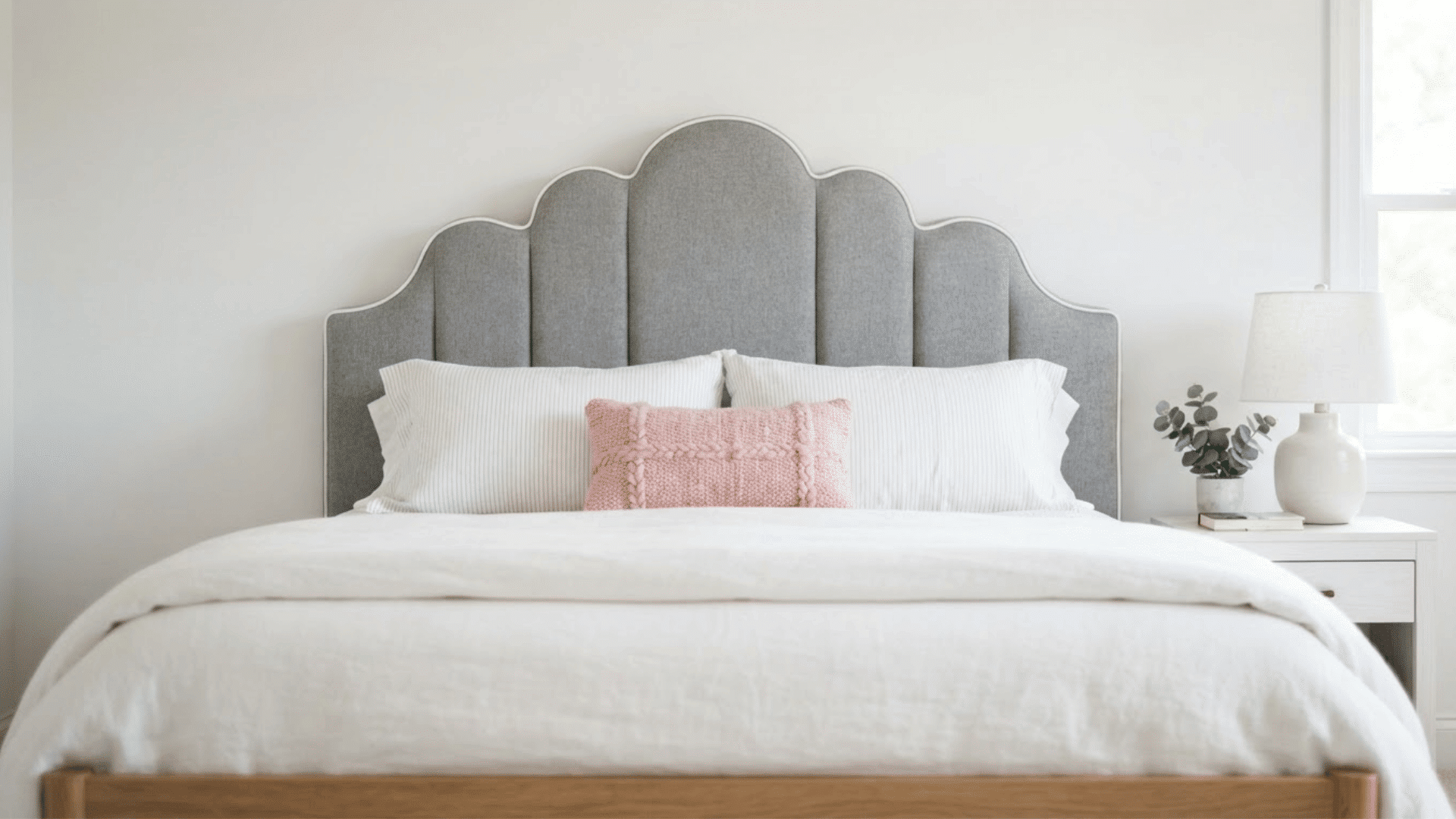 gray upholstered headboard with decorative piping and scalloped edges above a neatly made bed in a bright bedroom