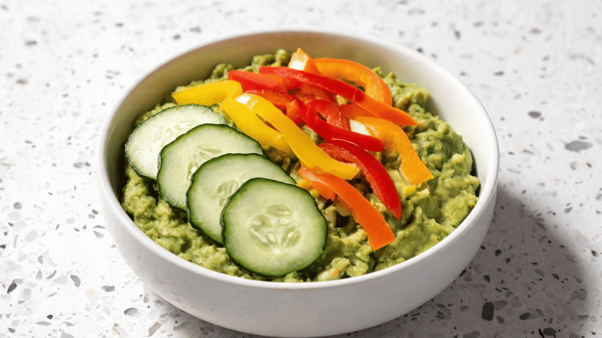 guacamole topped with cucumber slices and colorful bell pepper strips in a white bowl