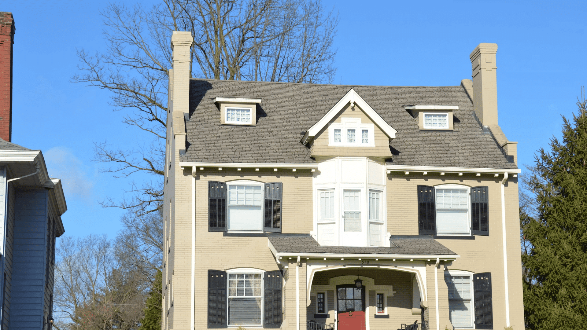 large beige brick house with black shutters red door and trees surrounding the property