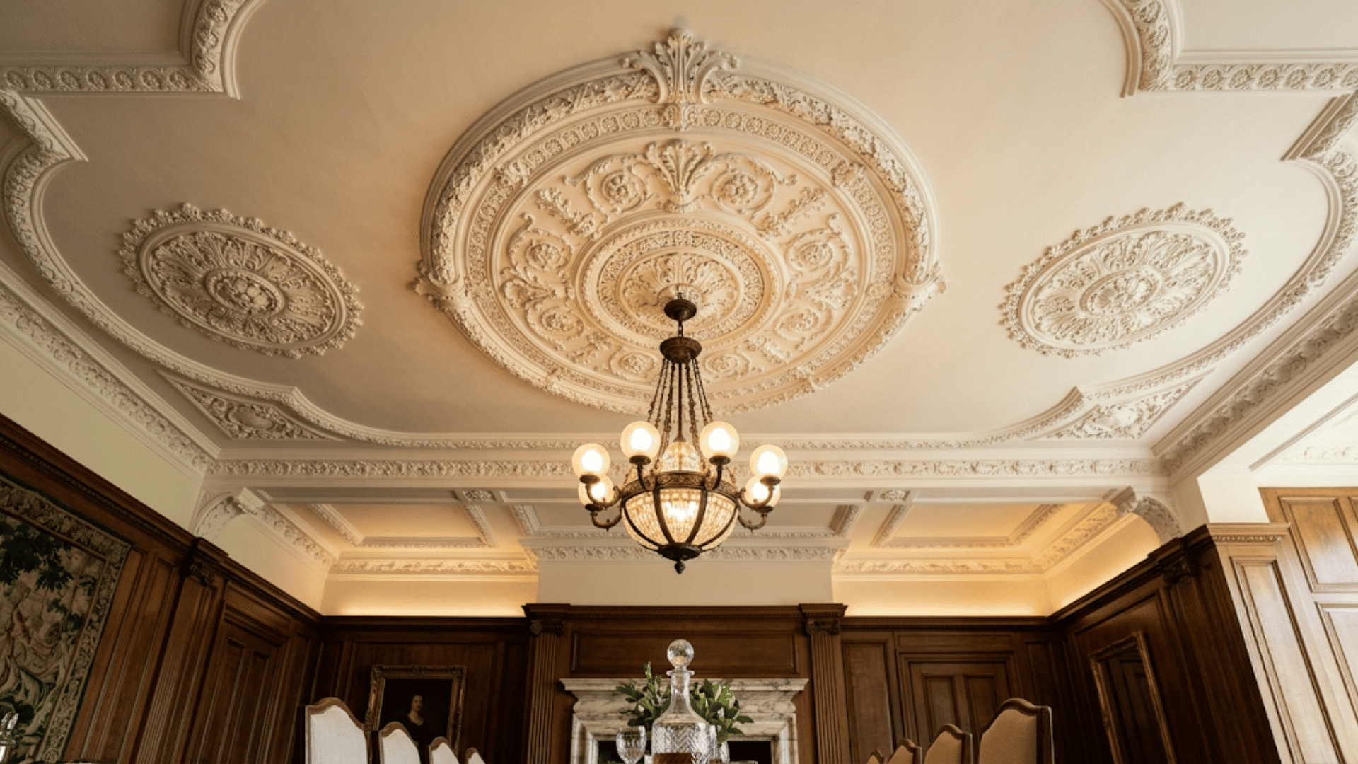ornate ceiling with decorative plaster details and classic chandelier above a formal room with dark wood paneling and fireplace (1)