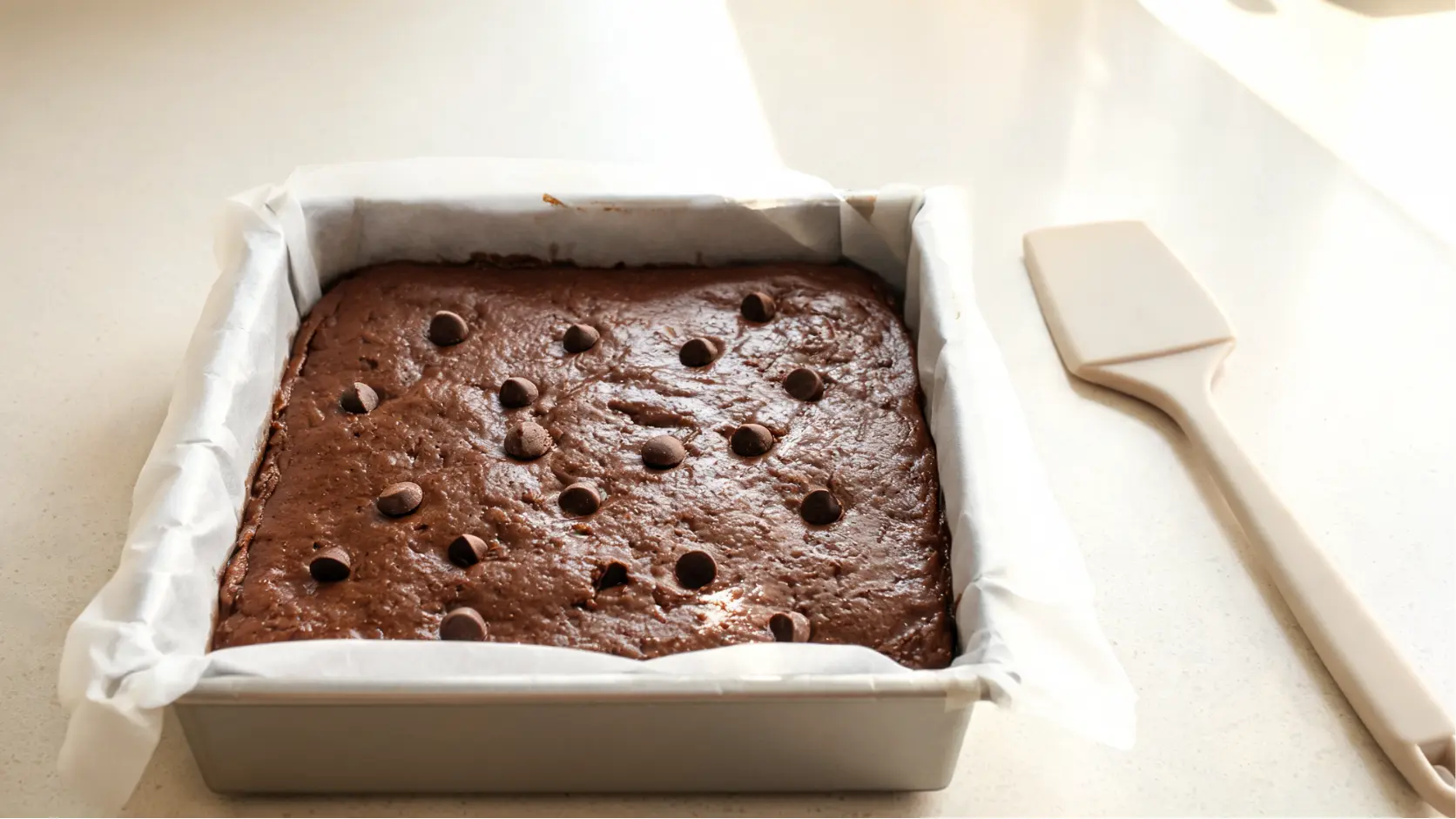paleo brownies batter spread in a parchment lined square pan with dark chocolate chips on top