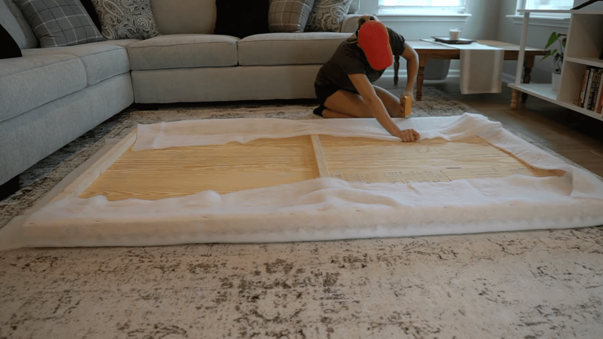 person stretching upholstery fabric over a wooden frame while securing it with a staple gun on a living room floor