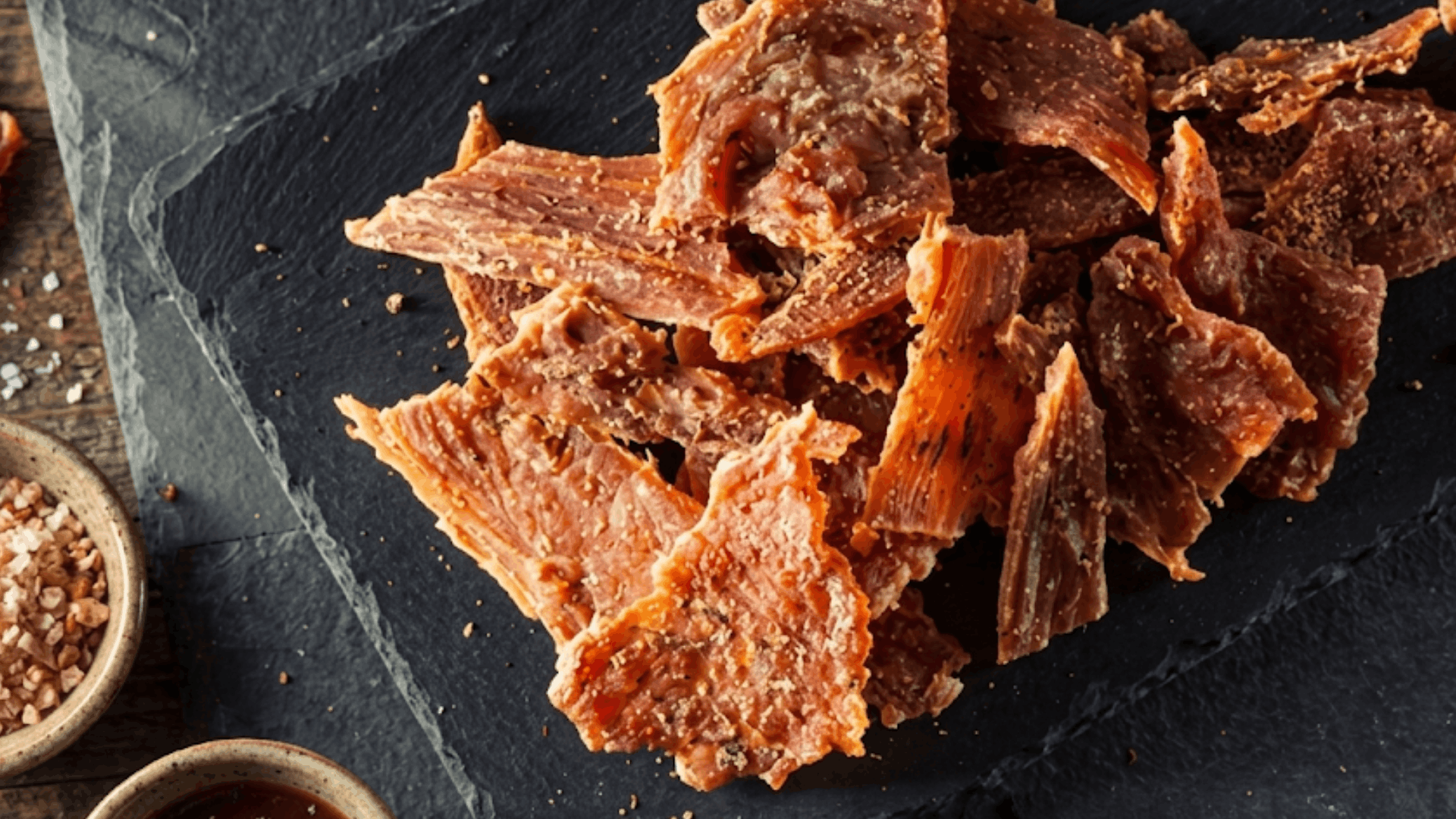 pieces of seasoned beef jerky arranged on a black serving board with coarse salt and spices for a high protein savory snack (1)