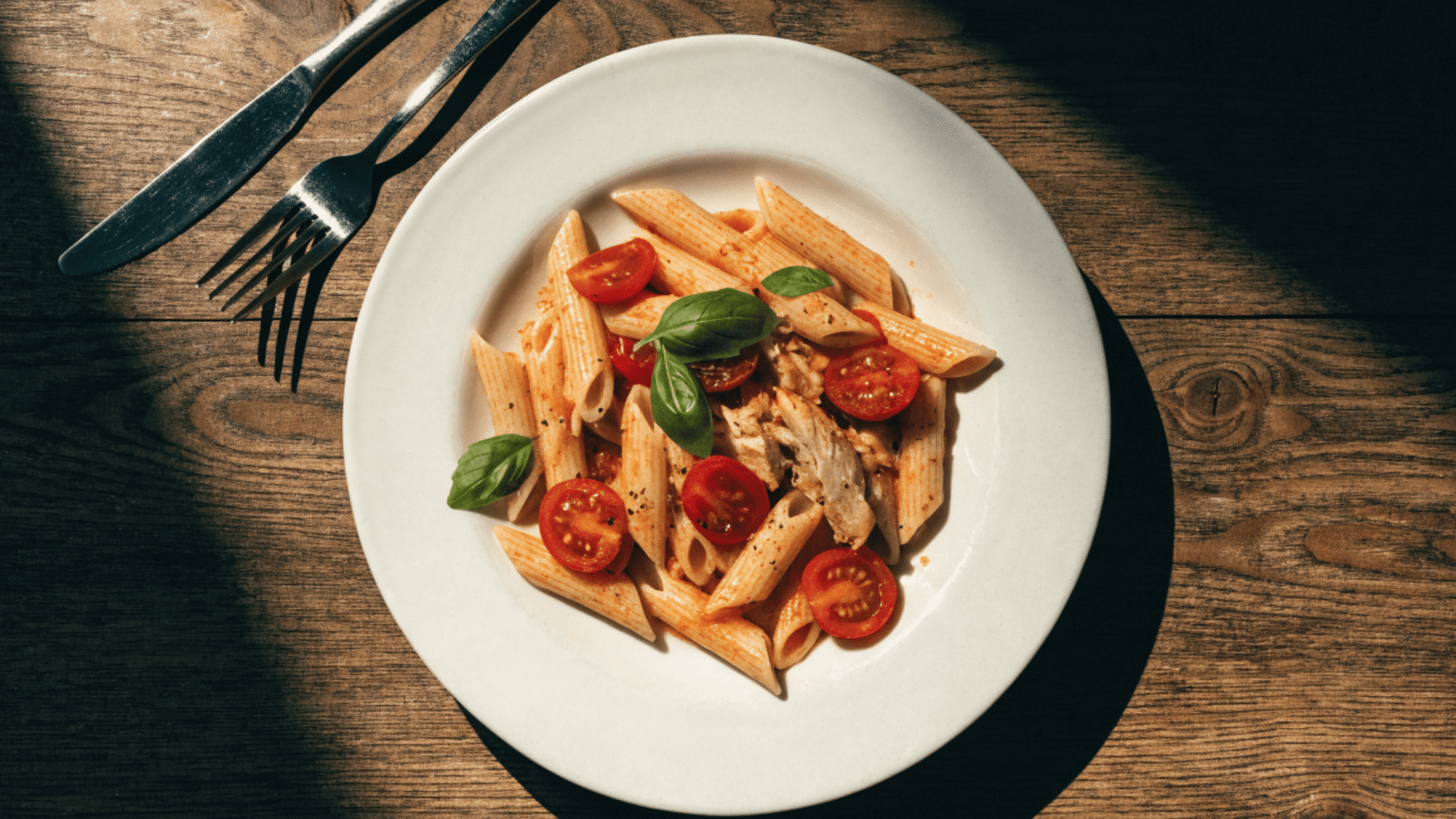 plate of penne pasta with tomatoes basil and chicken on a white dish beside fork and knife on wooden table
