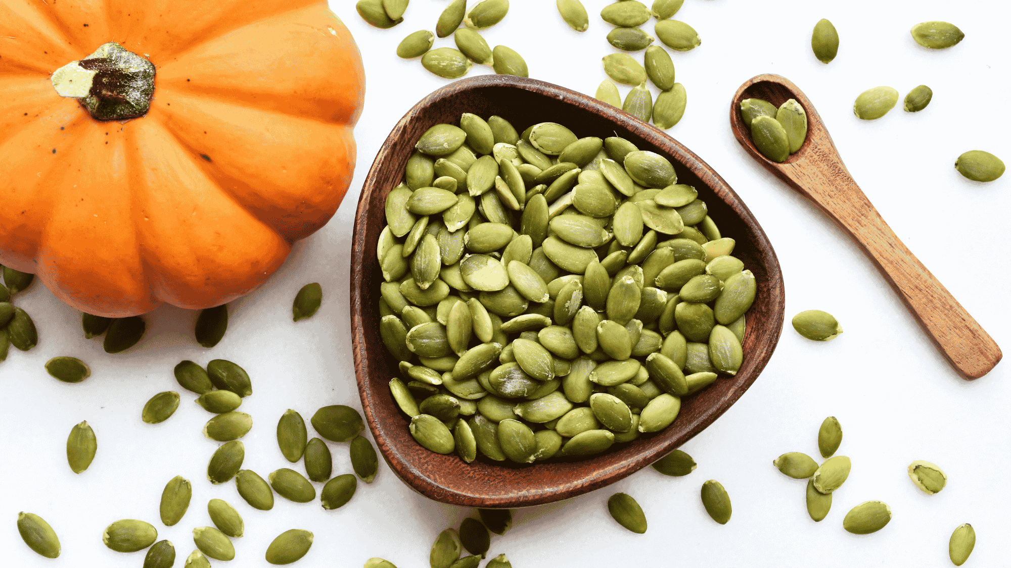 pumpkin seeds in a wooden bowl with fresh pumpkin pieces scattered on white surface