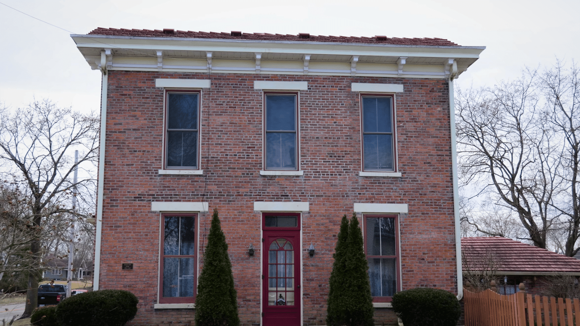red brick house with white trim and red door surrounded by shrubs and leafless trees on a cloudy day