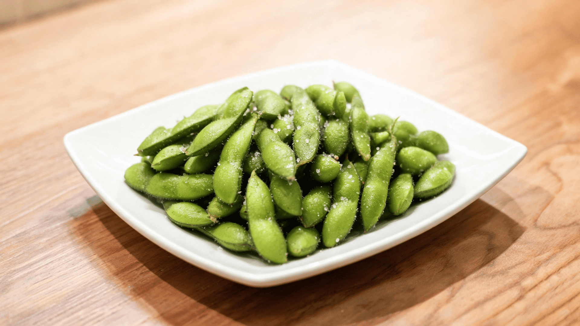 salted edamame pods served on a white square plate over a wooden table