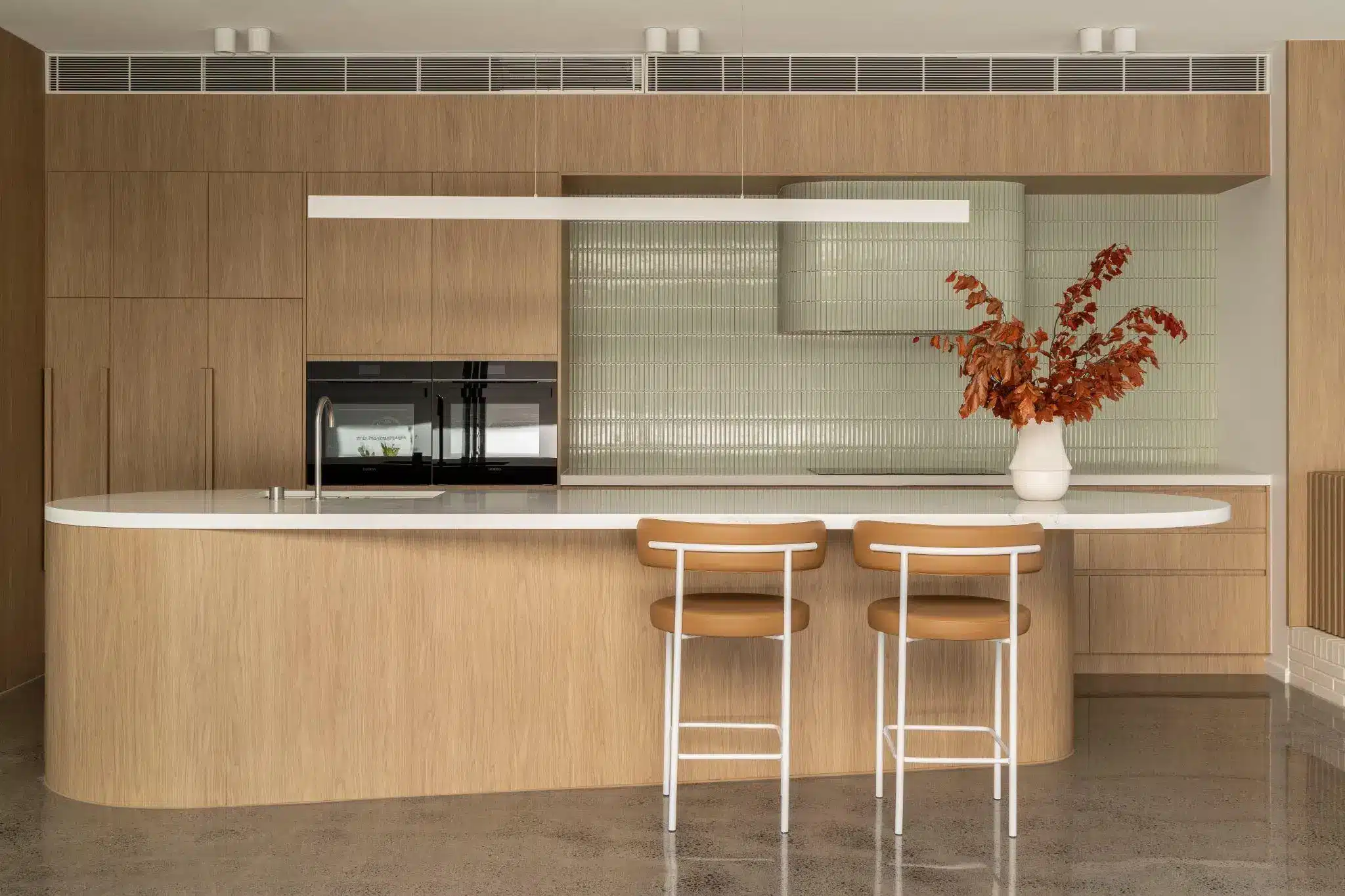 Modern kitchen with wood cabinetry, island, white countertop, and vase with dried orange leaves