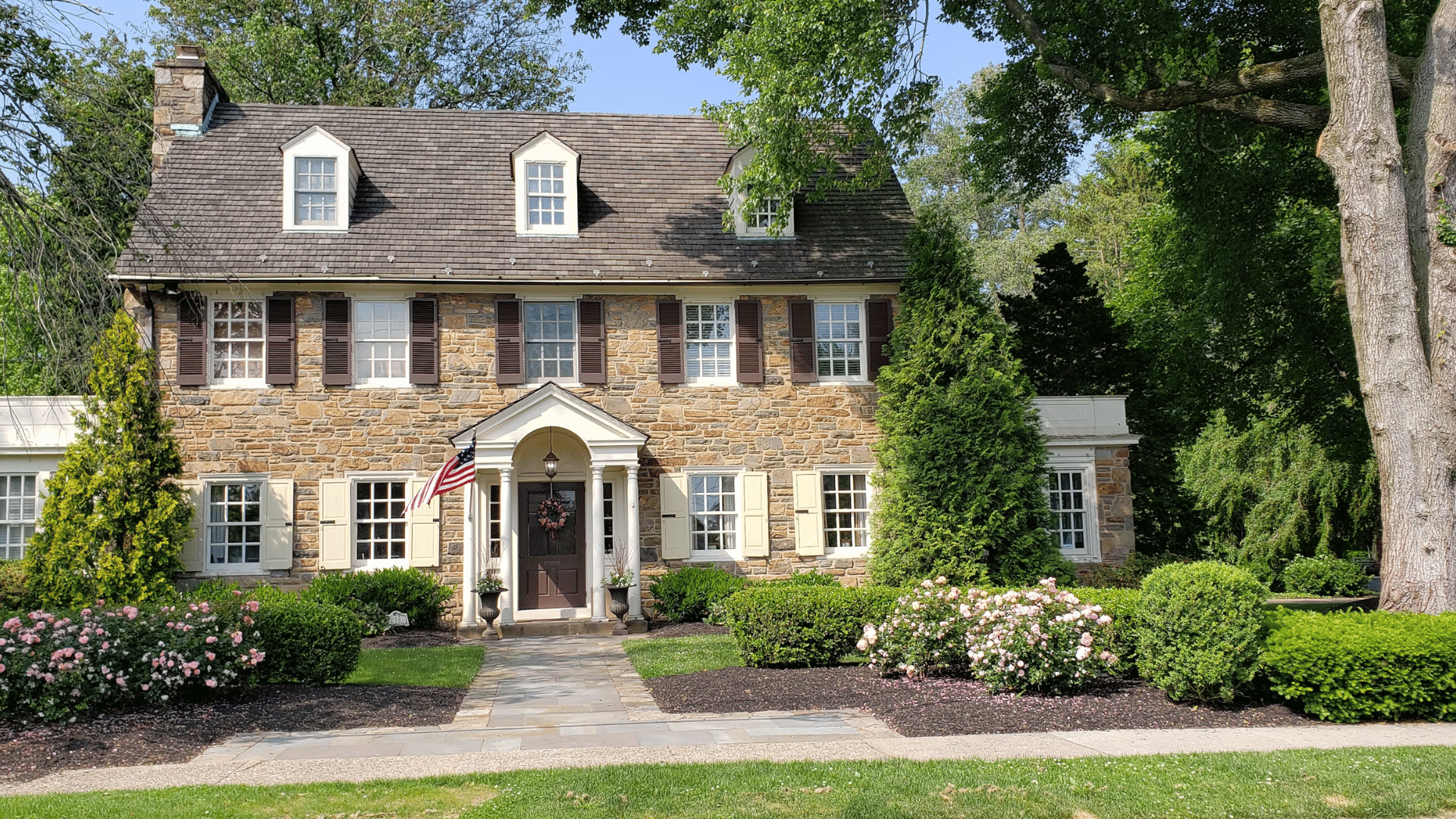 stone house with shutters front porch american flag and landscaped garden in bright daylight