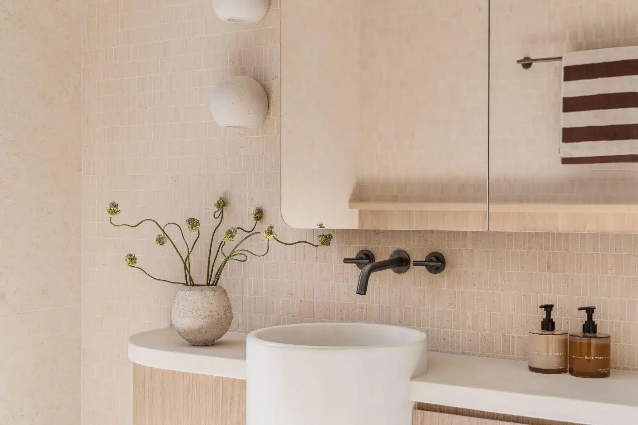 Modern bathroom with beige tile walls, ceramic vase with flowers, and black faucet above round sink