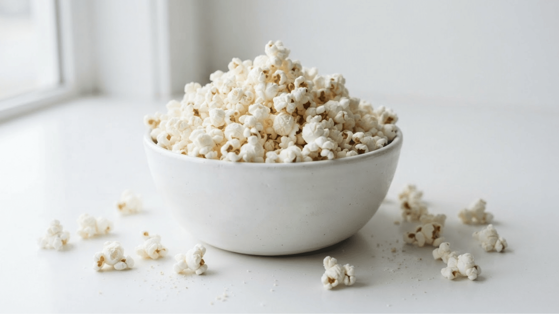 white ceramic bowl filled with freshly air popped popcorn with loose kernels scattered on a bright windowsill