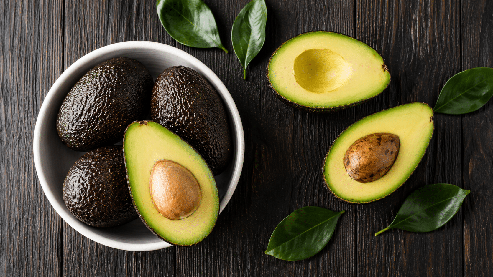 whole and halved avocados with creamy green flesh arranged in a white bowl on a dark wooden surface with fresh green leaves