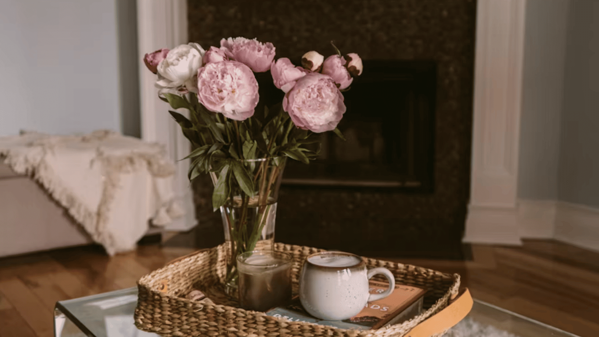 woven tray with pink flowers coffee mug and candle arranged on a glass coffee table near a fireplace