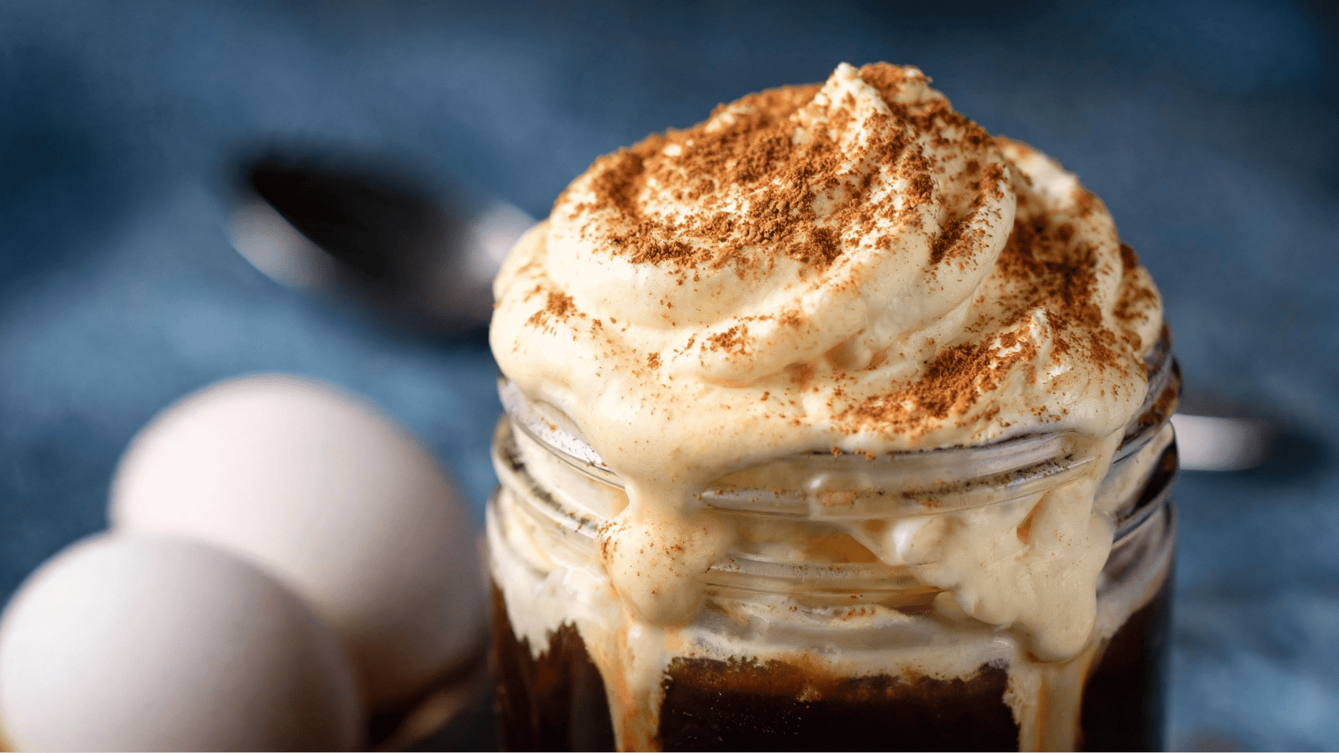 Creamy egg coffee topped with whipped foam and cocoa powder served in a glass with eggs beside it.