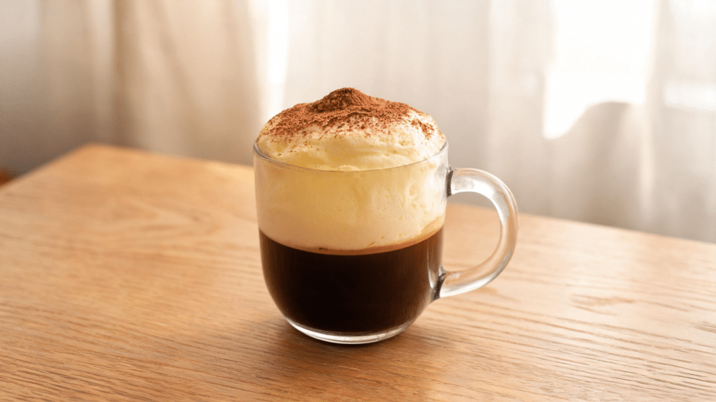 Creamy egg coffee with whipped foam topping and cocoa powder served in a glass cup on a wooden table.