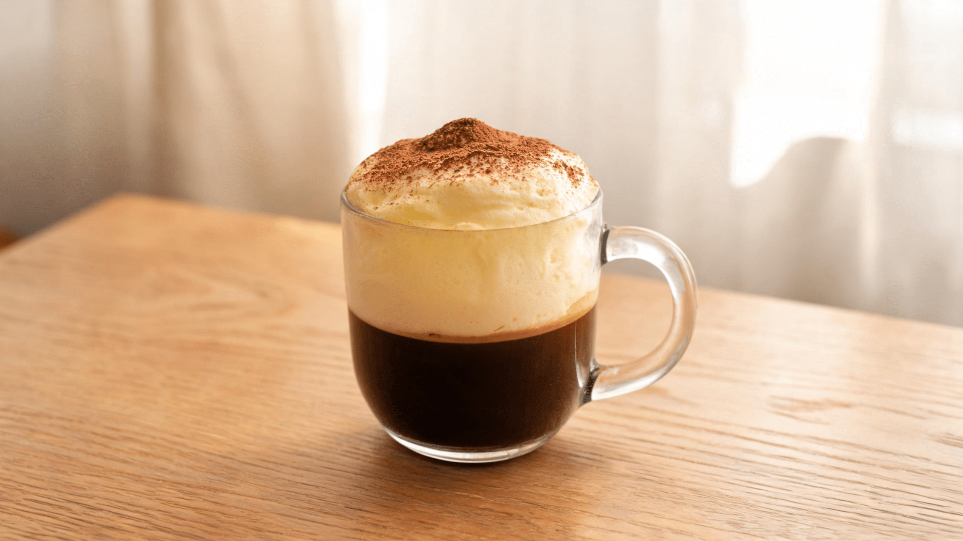 Creamy egg coffee with whipped foam topping and cocoa powder served in a glass cup on a wooden table.
