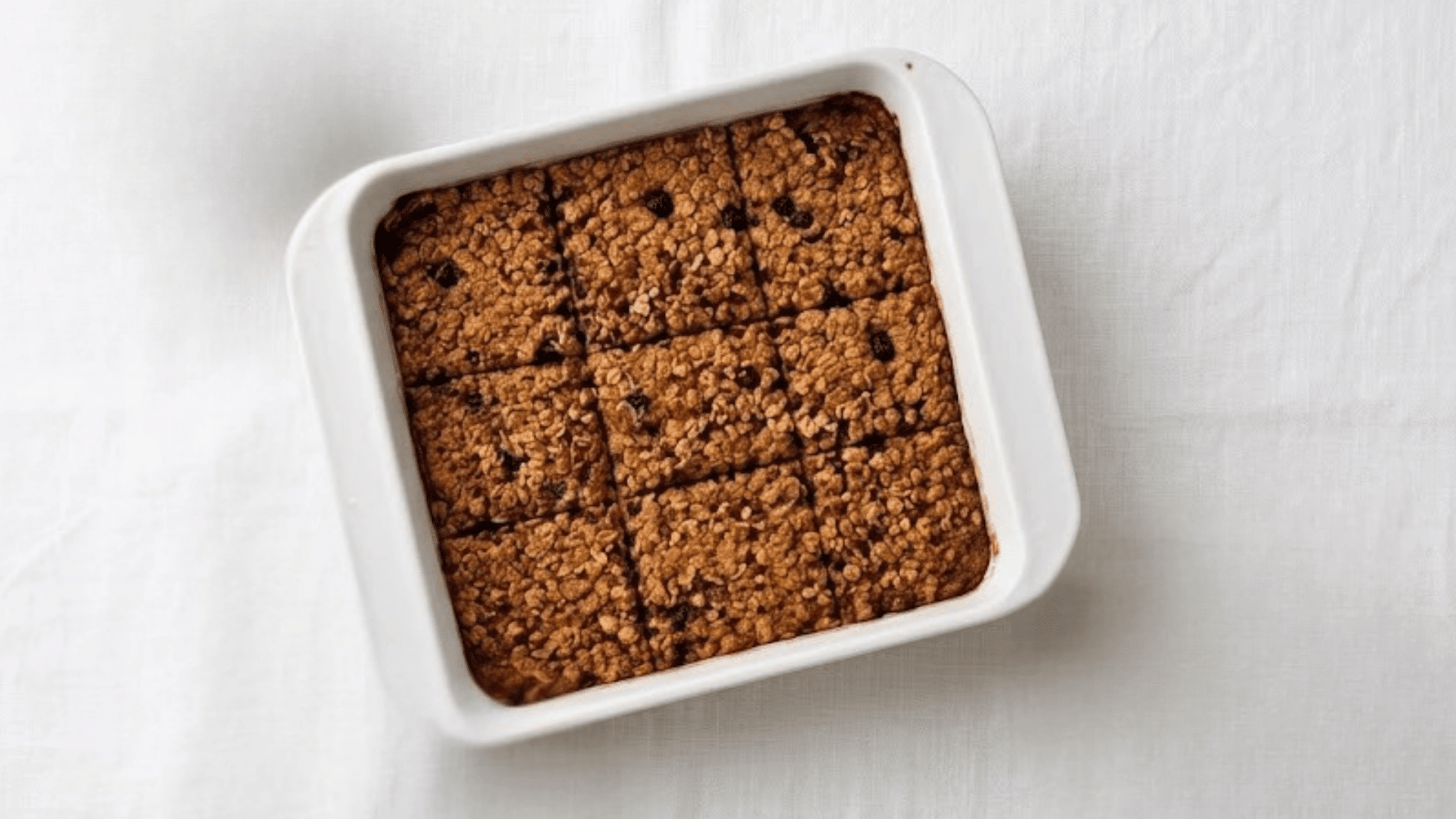 a baking dish of baked oatmeal cut into squares on a white table with a bowl of yogurt granola and a folded cloth nearby