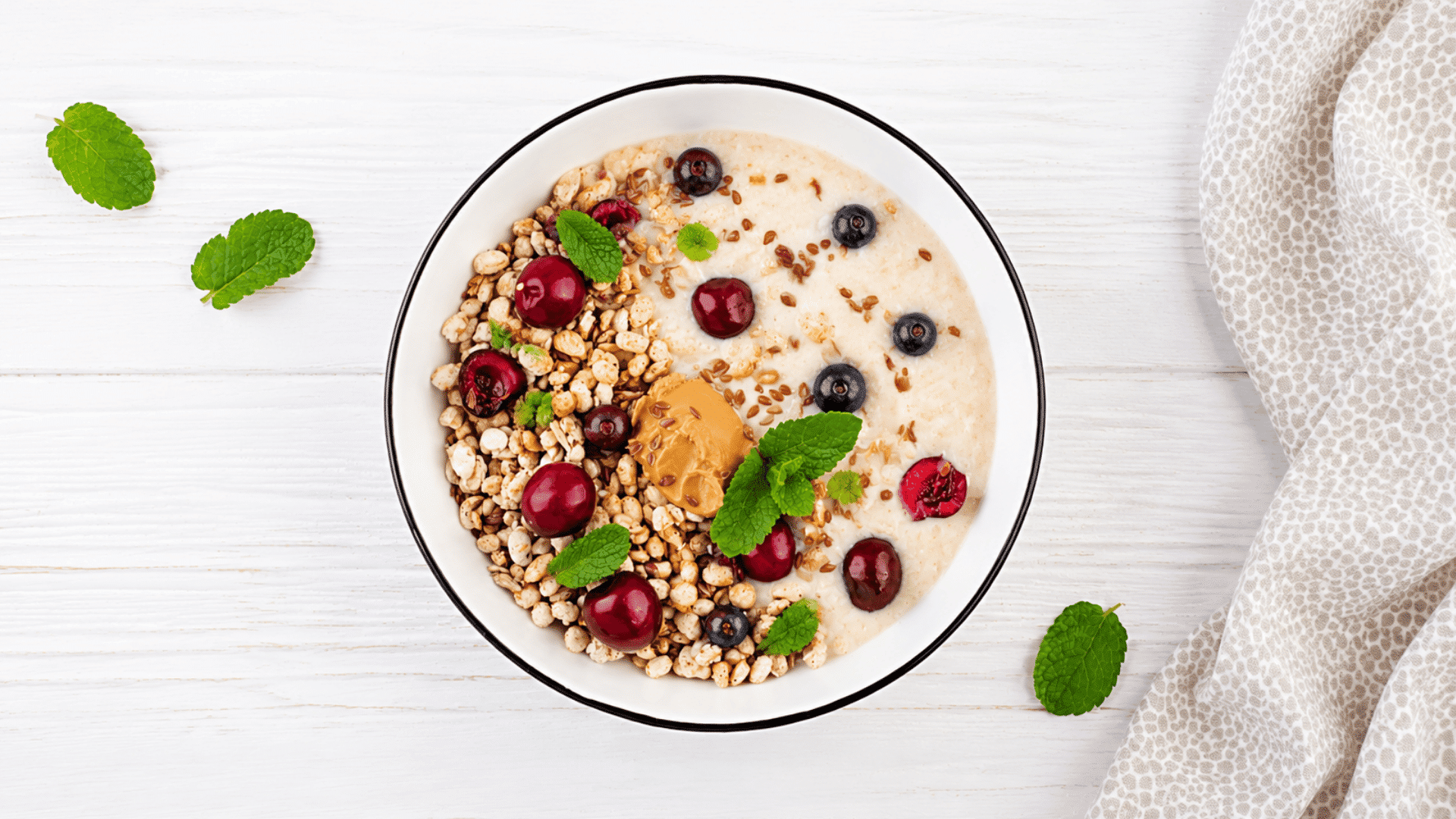 a bowl of creamy oatmeal topped with granola peanut butter fresh cherries blueberries flaxseeds and mint leaves on a white table