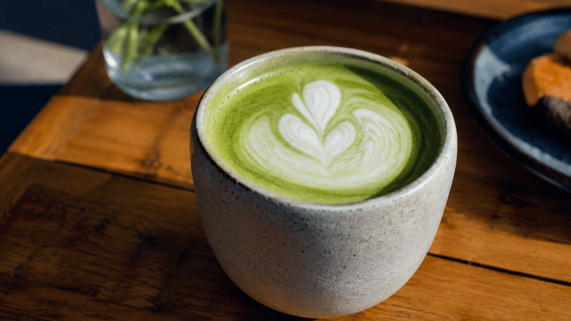a ceramic cup of creamy matcha latte with heart shaped latte art resting on a wooden table in a cozy cafe setting